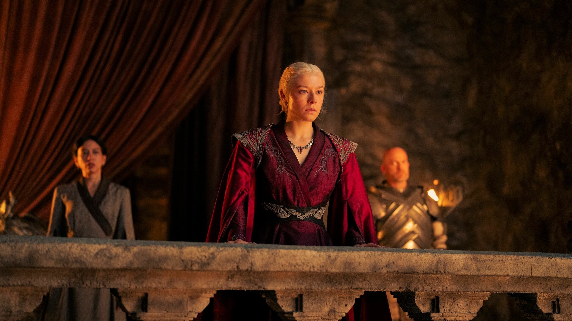 A woman in regal dress stands on a balcony in a cavern.