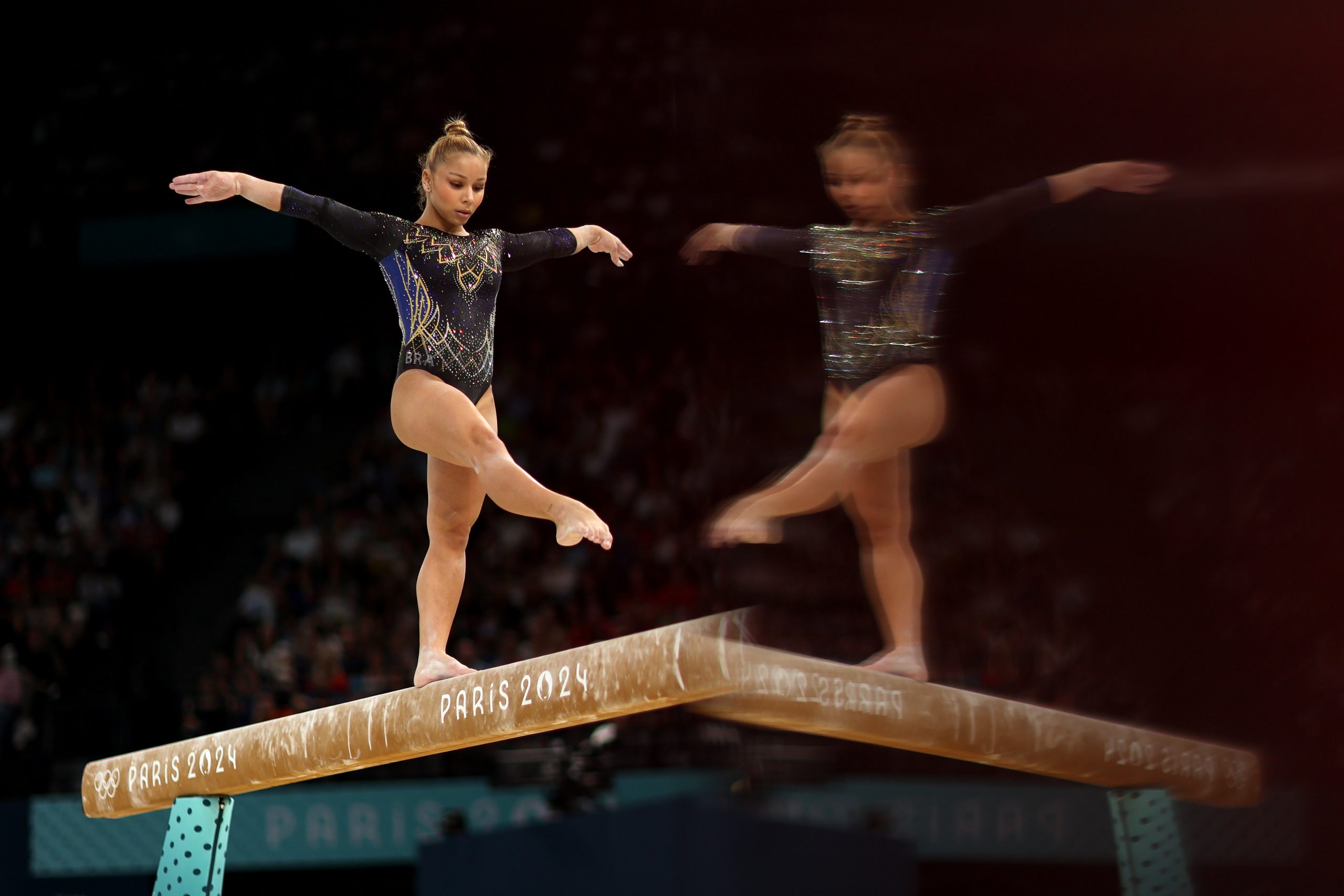 A mirrored image of Saraiva balancing with one leg up on the beam. 