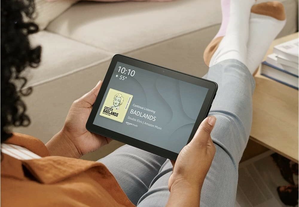 a person sits with legs extended on a coffee table while holding the fire HD 8 tablet in their hands. The display screen of the tablet shows the time, temperature, and the current song playing