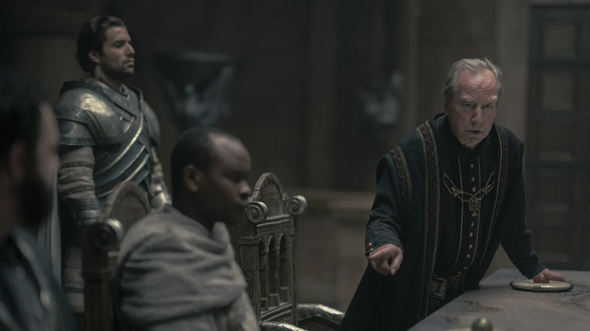 Men in medieval clothing sit and stand around a table in a castle room.