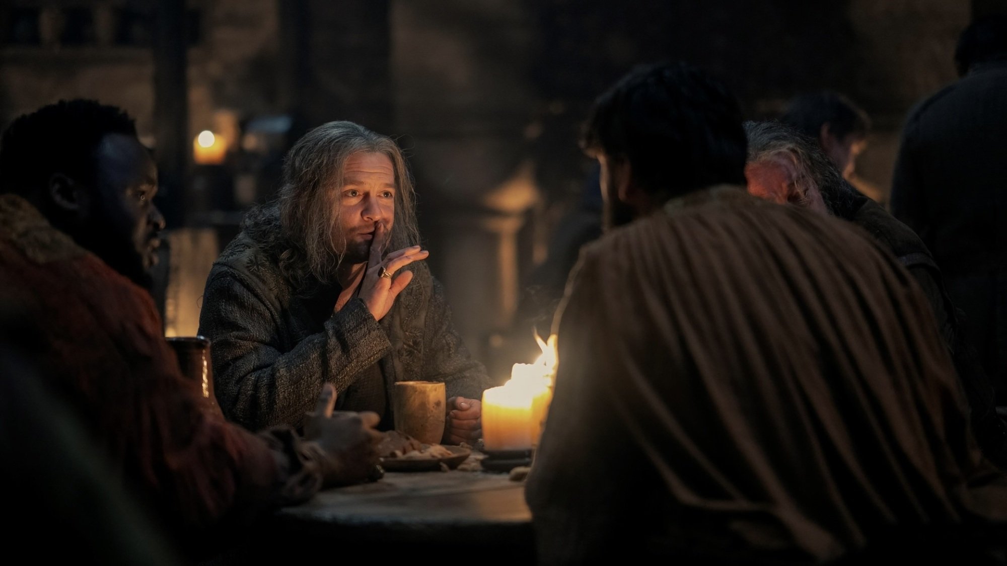 Ulf the White sits at a candlelit table in a tavern, holding a finger over his lips as he tells people around him a story.