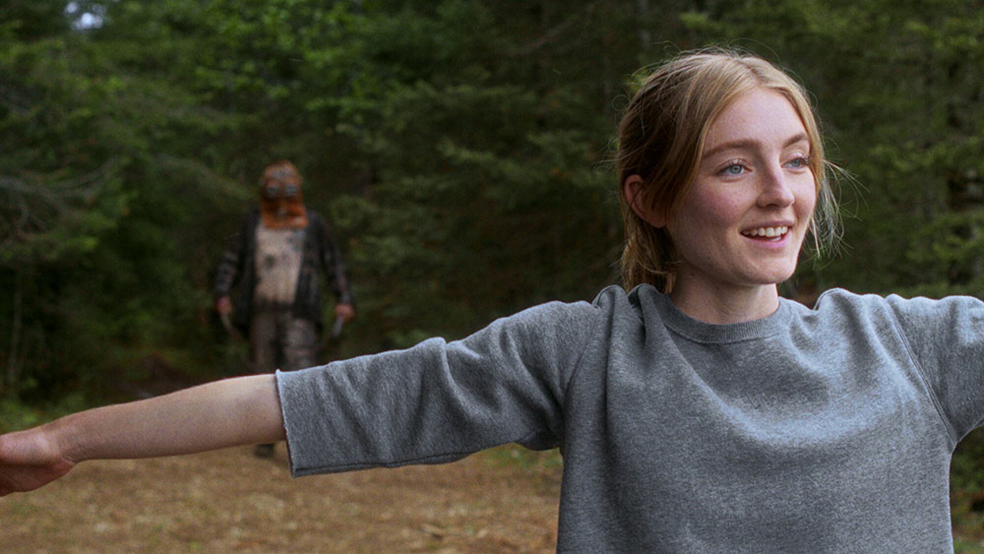 A woman stands smiling, facing away from a masked man who emerges from the woods behind her.