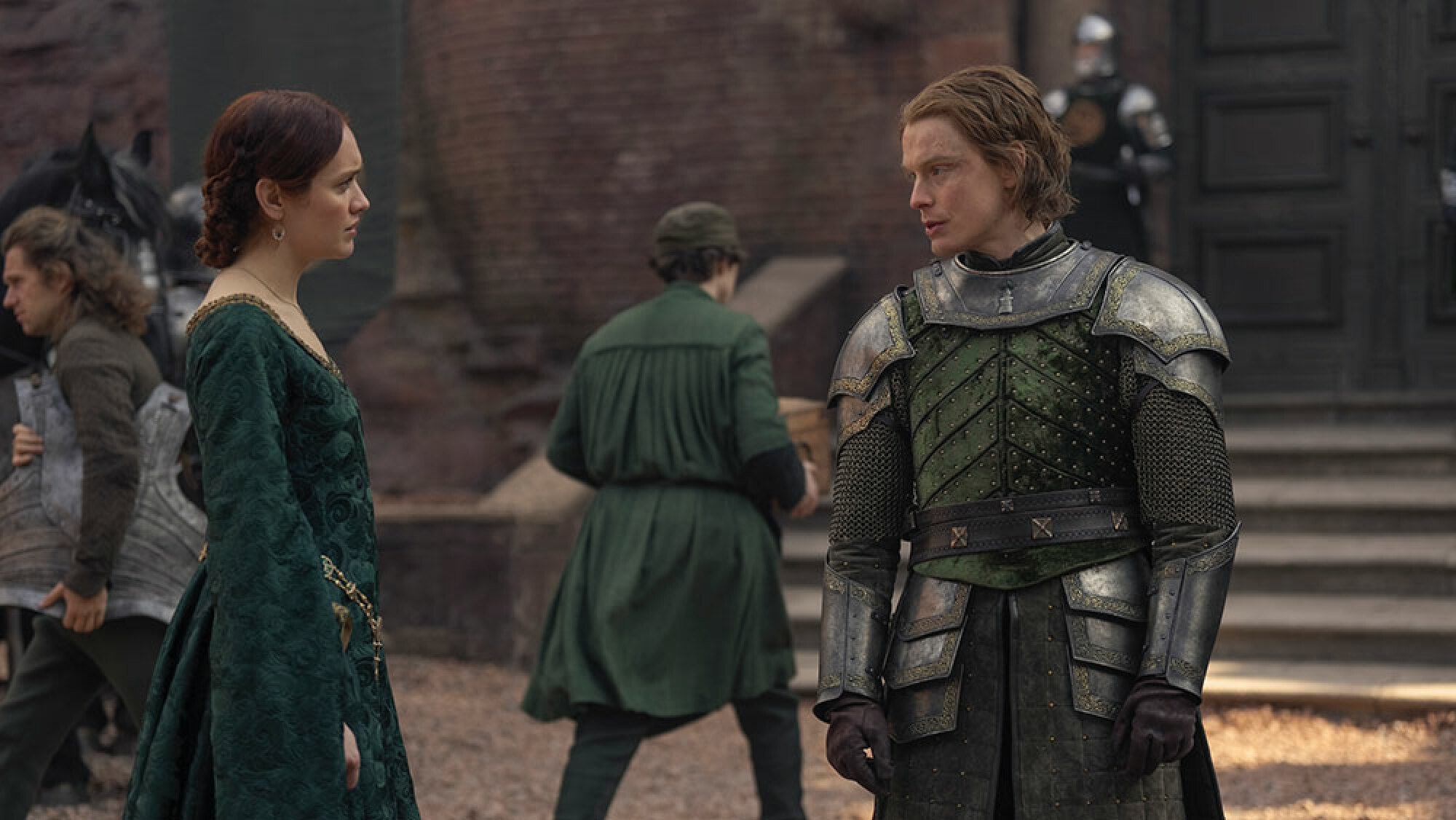 A woman in regal medieval clothes talks to a man in armour in a courtyard.