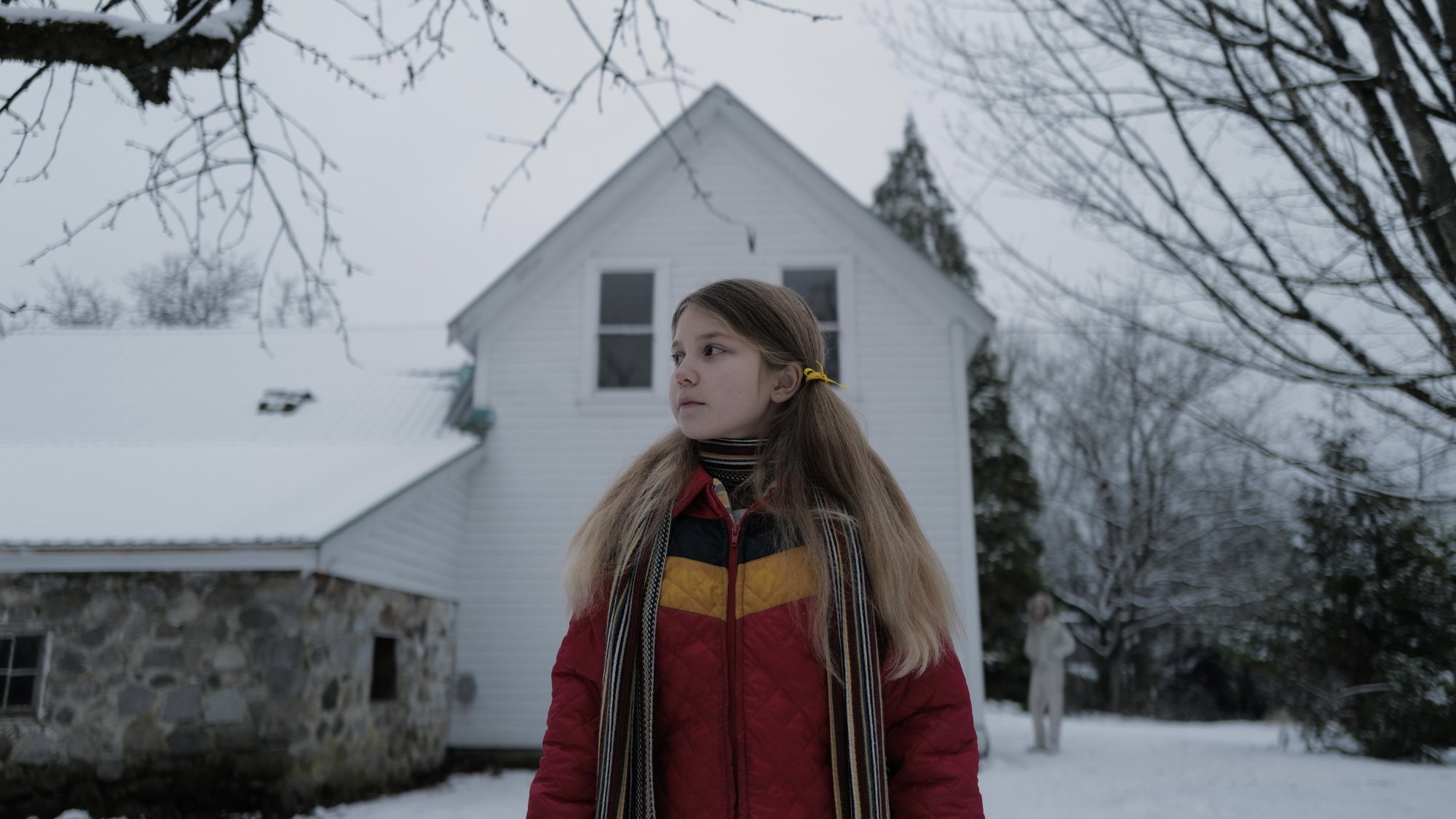 A young girl in a colorful winter coat stands outside her white house in the snow, while a man lurks deep in the background.