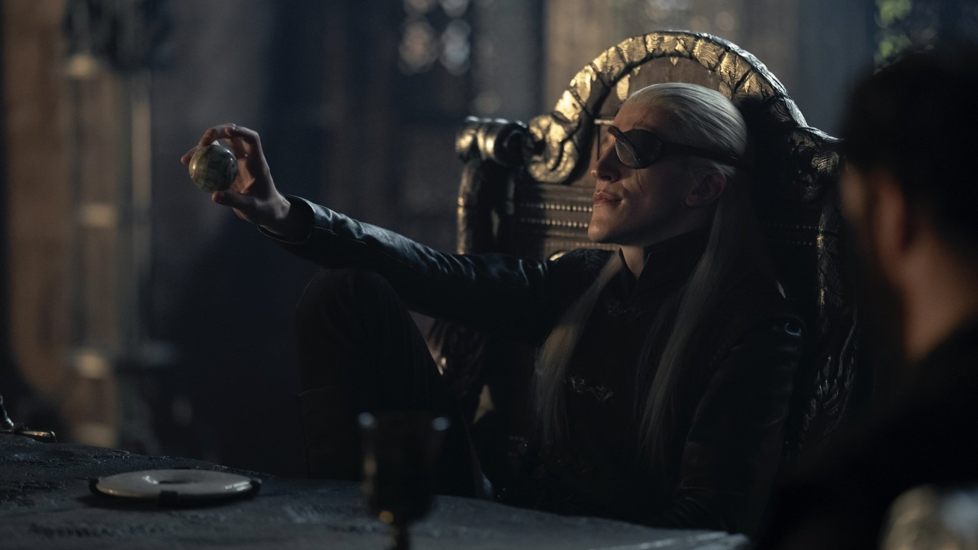 Aemond Targaryen sits at the head of his Small Council table, holding up a marble ball.