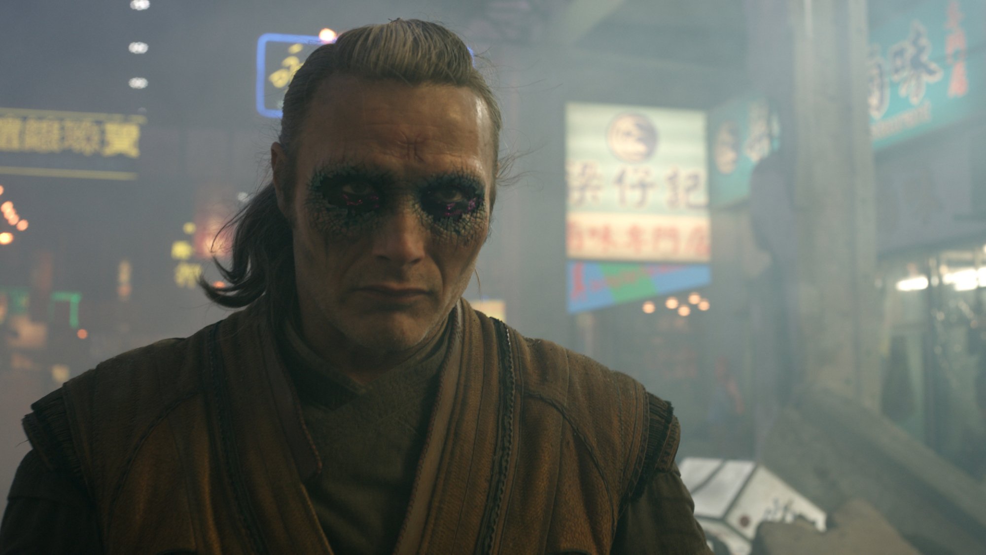 A man with dark makeup around his eyes and a low, short ponytail, wearing robes. In the background, the city of Hong Kong is under attack.