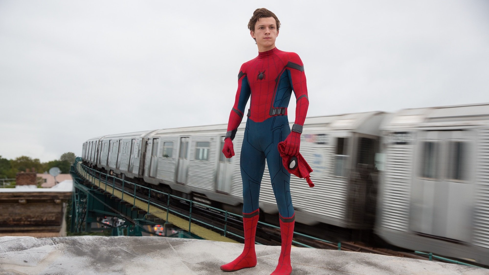 Tom Holland as Peter Parker, standing on a roof and wearing the Spider-Man suit, except for the mask, which he holds in his hand. A blurry New York City subway train speeds by in the background.