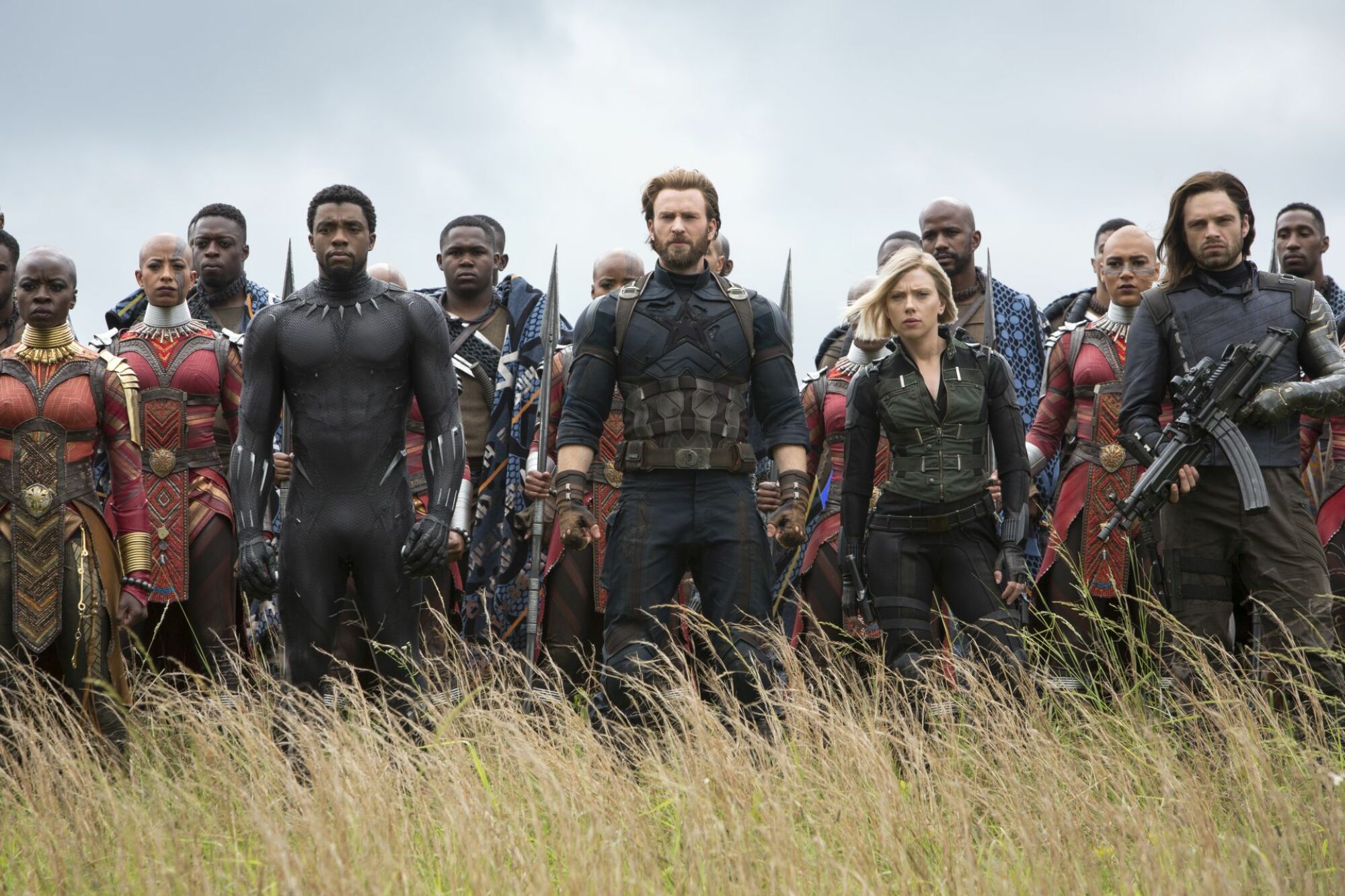 Wide shot of Captain America (Steve Rogers) in a dark, rugged super soldier suit, leading battle forces including soldiers from Wakanda, Bucky Barnes, and Black Widow. They assemble in a field, awaiting an attack.