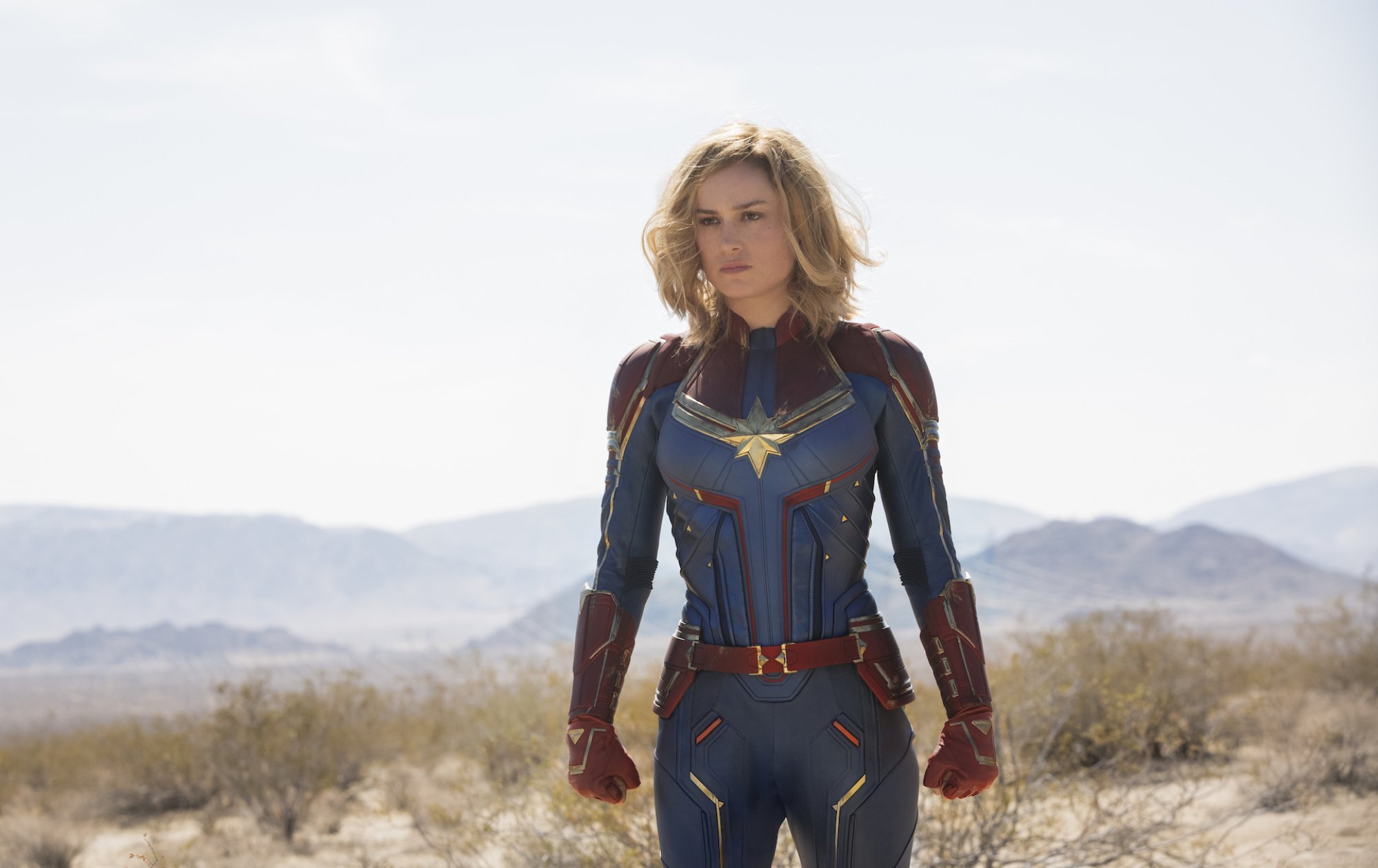 Carol (Brie Larson) standing in a field, wearing her red and blue Captain Marvel suit, with a distinct gold star over the chest.