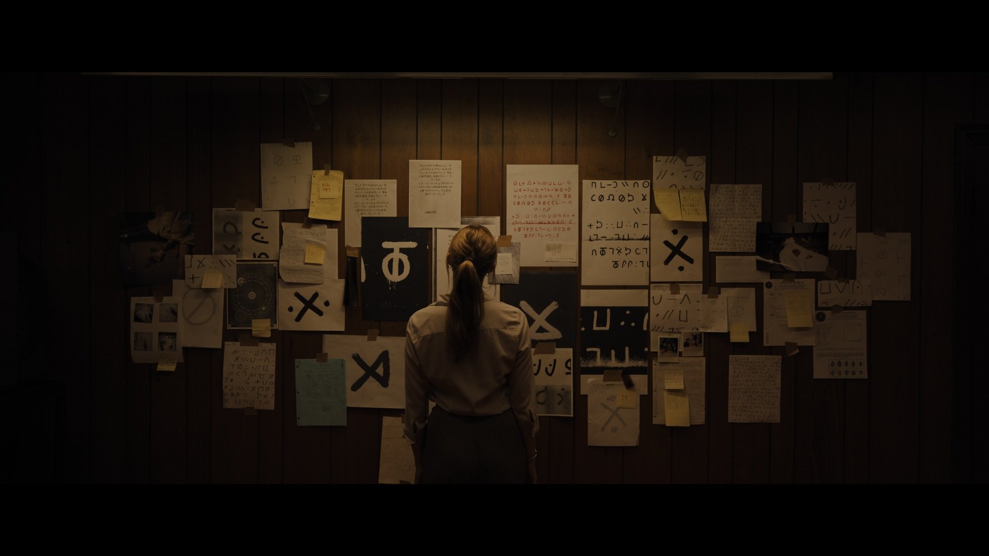 A young woman looks at a wall covered in cryptic messages and police reports.