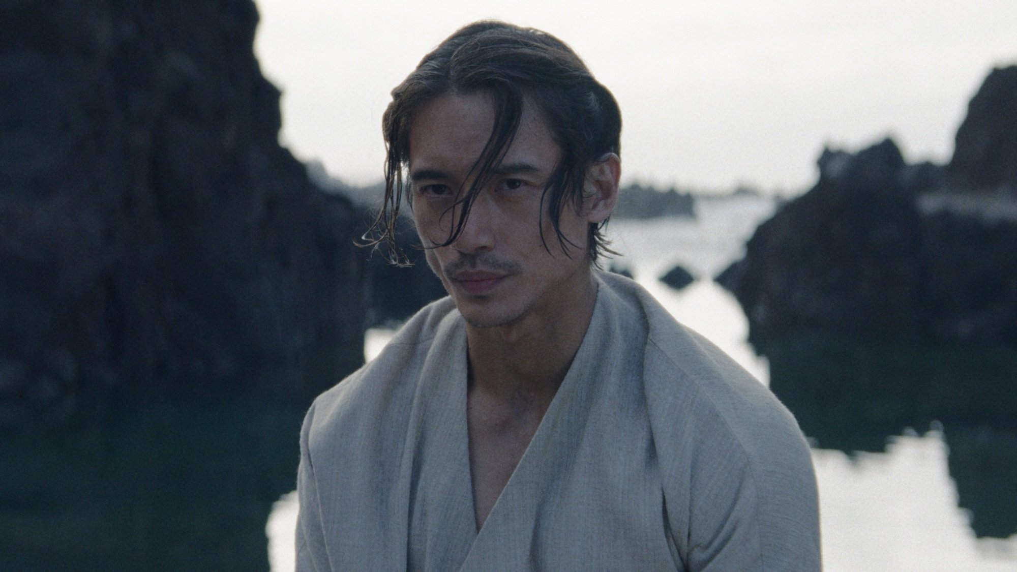 The Stranger stands with his back to the ocean, wearing a white shirt.