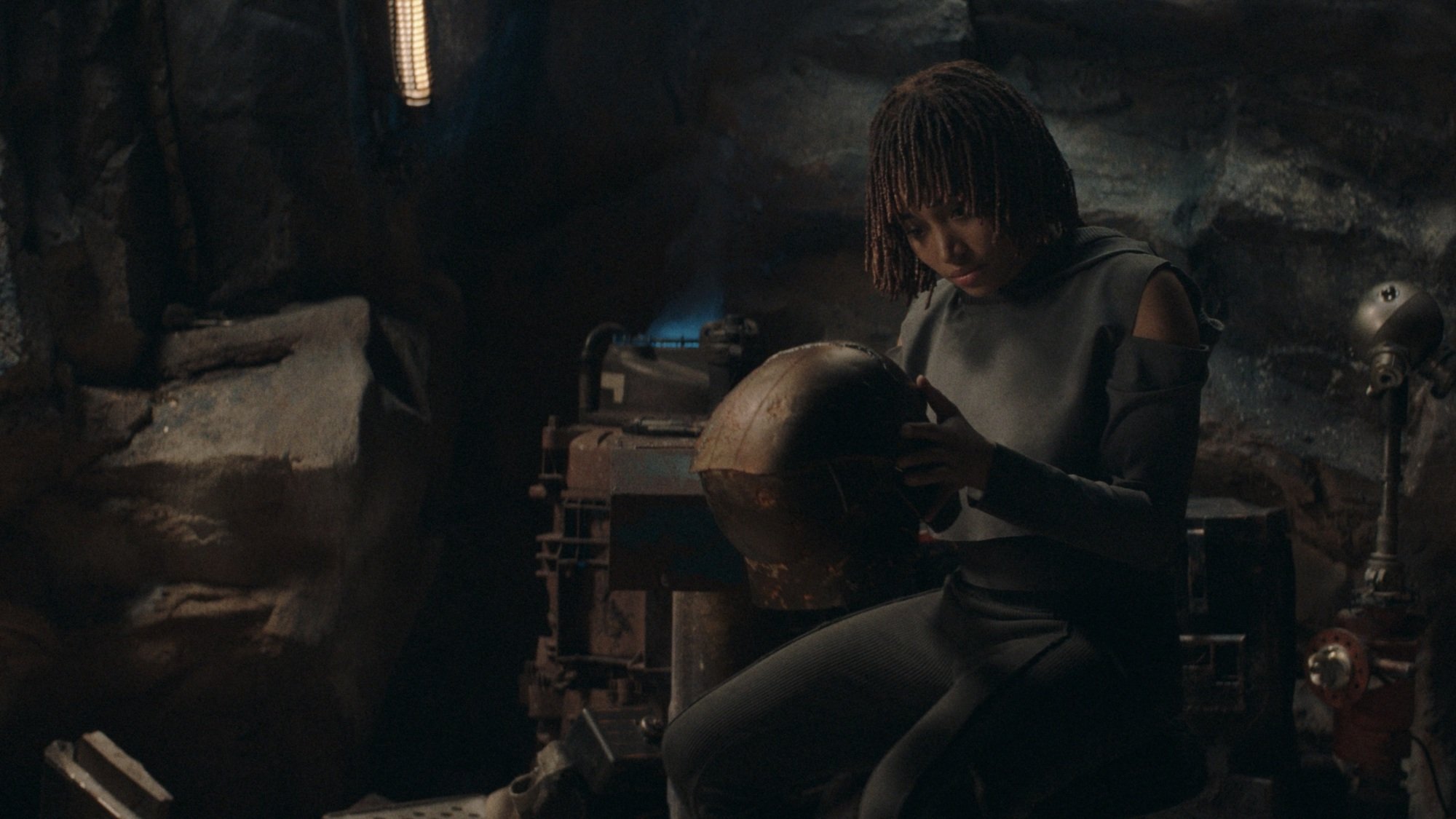 Osha sits in the Stranger's cave, examining his cortosis helmet.