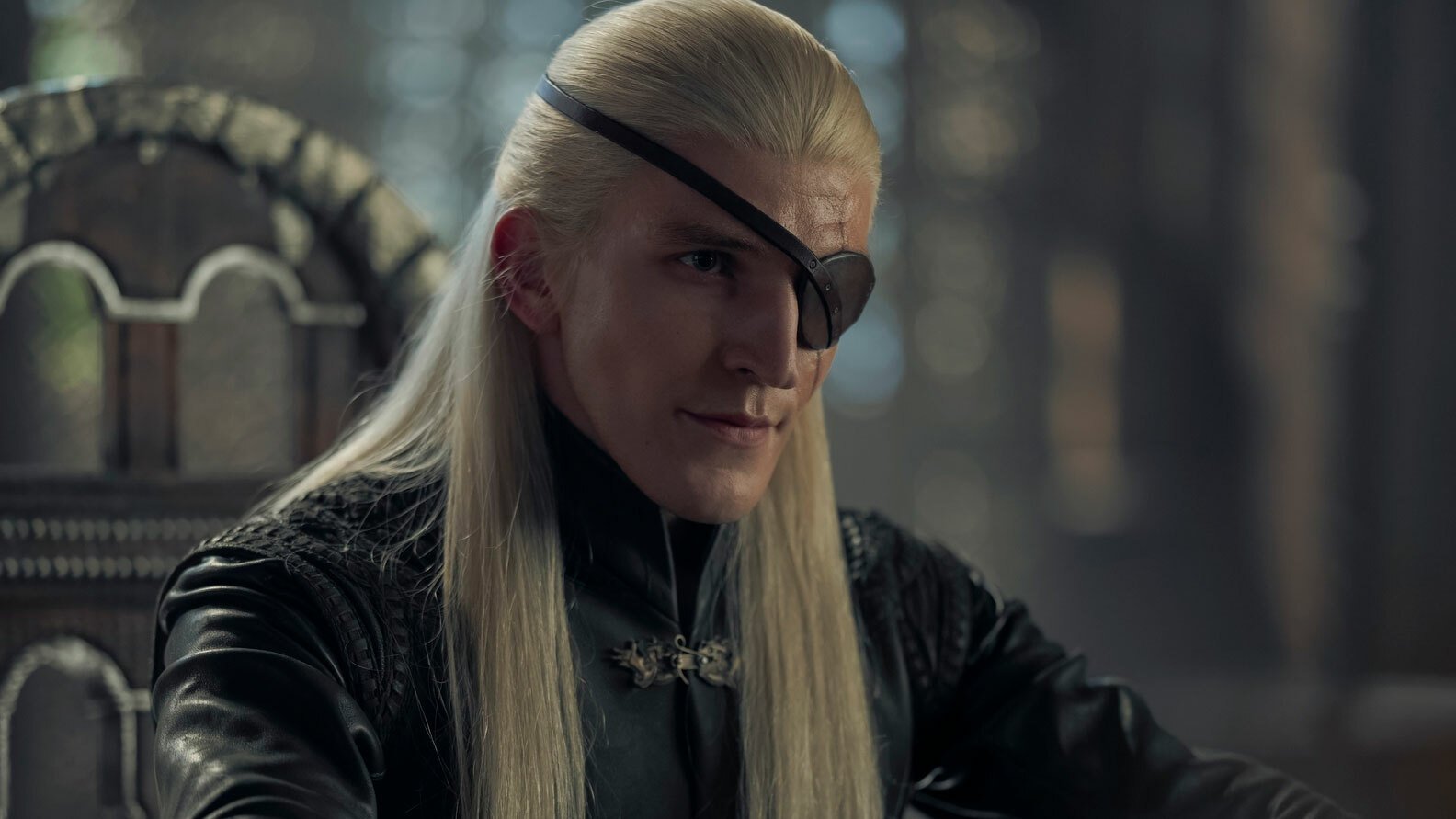 A man with long blonde hair and an eyepatch sits on a chair.