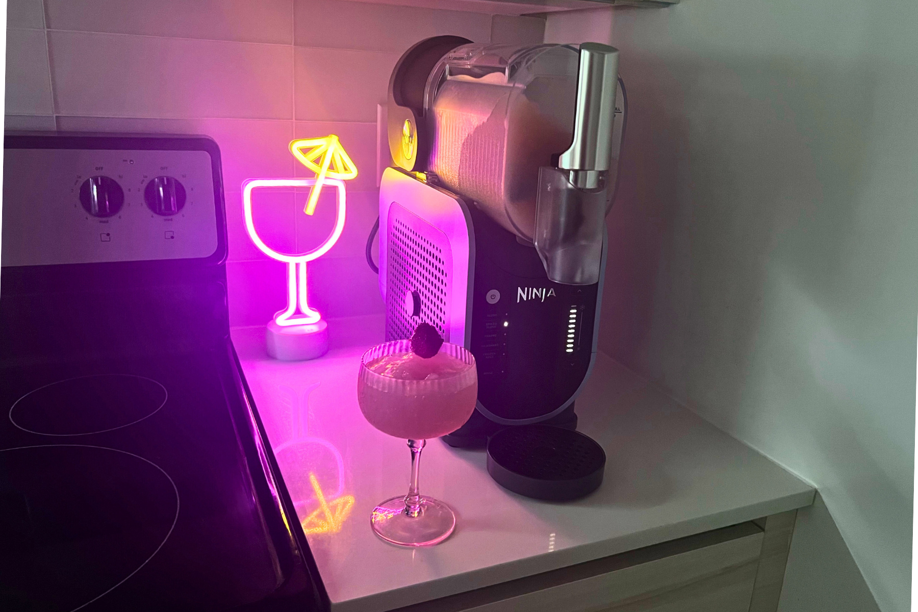 Glass of frosé on countertop beside Ninja Slushi machine with neon cocktail light in background