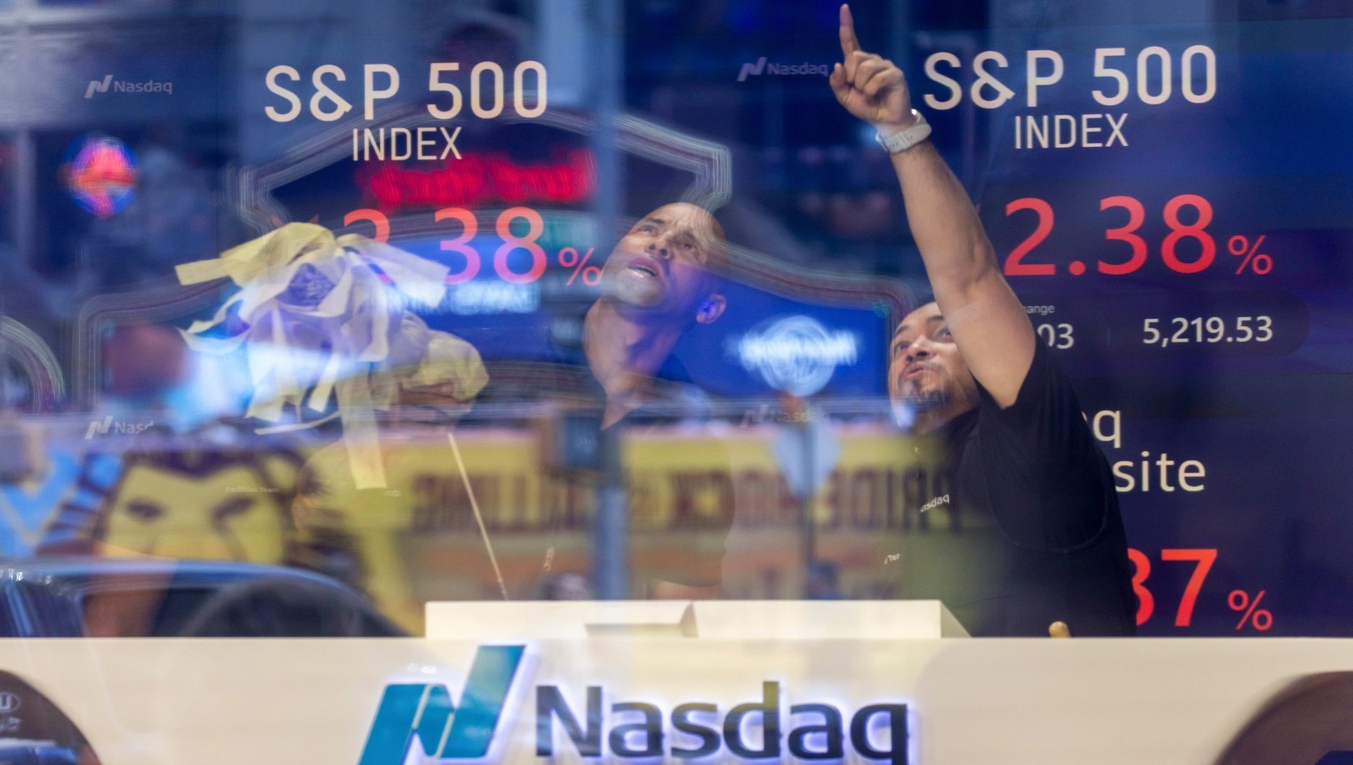 Two men at the NASDAQ stock exchange point at falling numbers on a screen. 