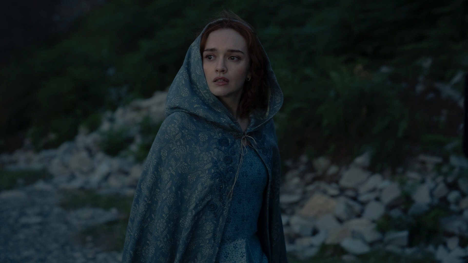 Alicent Hightower stands outside on the island of Dragonstone, wearing a blue dress and cloak.