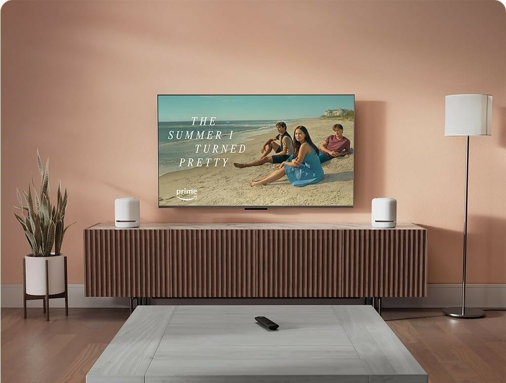 a living room setting with an Amazon fire tv mounted on the wall, amazon device speakers beside the tv and an Amazon fire TV stick remote on the ottoman.