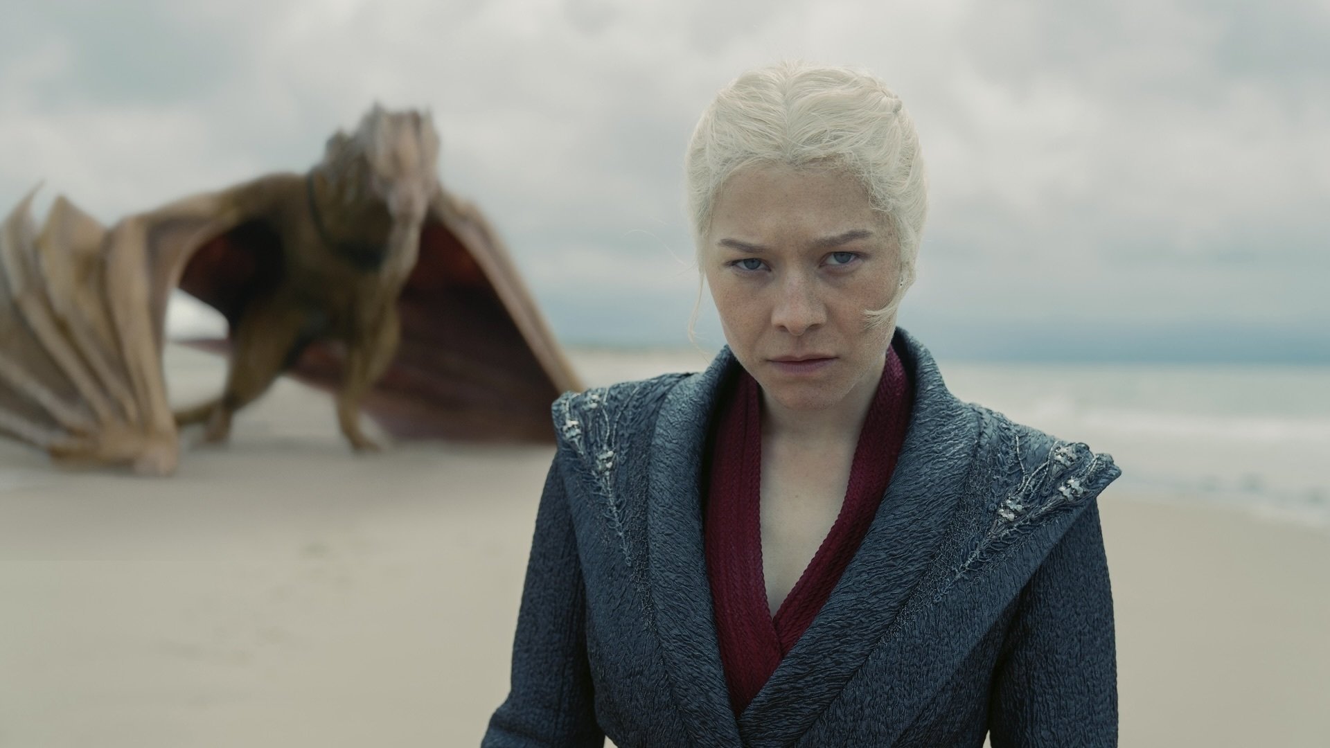 Rhaenyra Targaryen stands on a beach with her yellow dragon Syrax behind her.