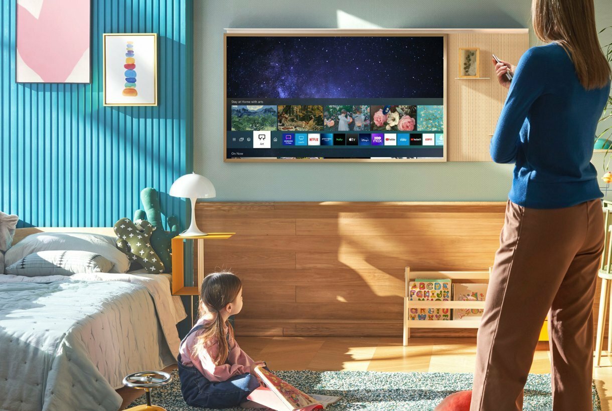 Parent pointing remote at Samsung The Frame TV hanging on wall and child sitting on floor near bedroom furniture in peripherals