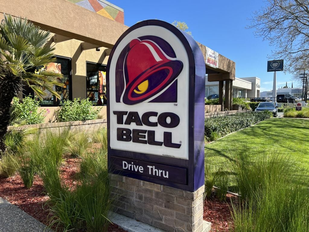 Taco Bell drive-thru open for business with lush green plants and clear sky in a residential neighborhood