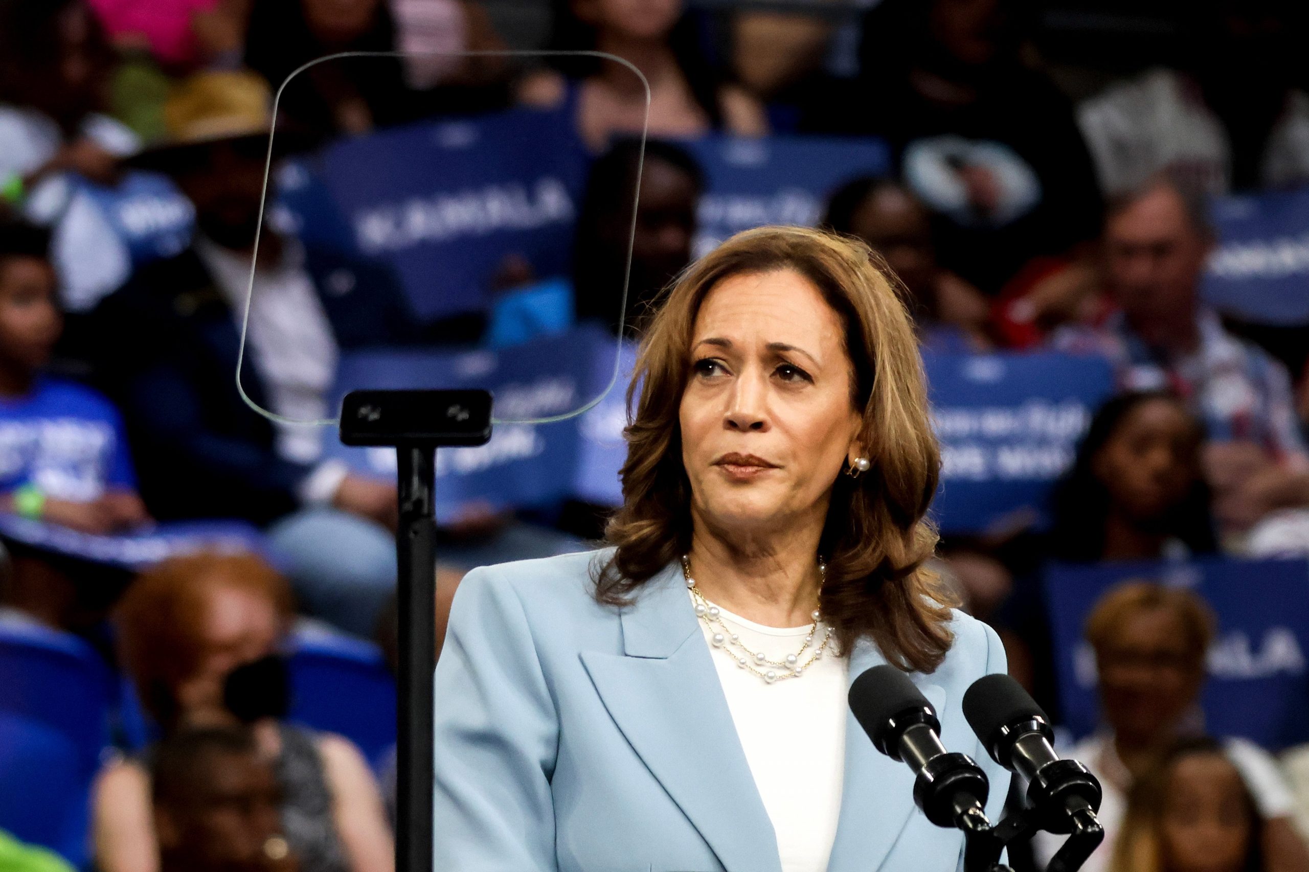 Vice President Kamala Harris looking serious in a pale blue suit at a campaign rally.