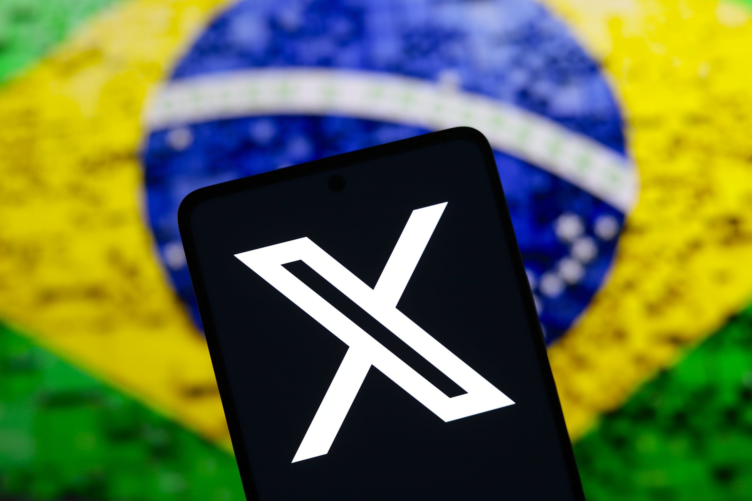 The X (Twitter) logo is displayed on a smartphone screen with a flag of Brazil in the background. Social network X has announced that it will close its office in Brazil.