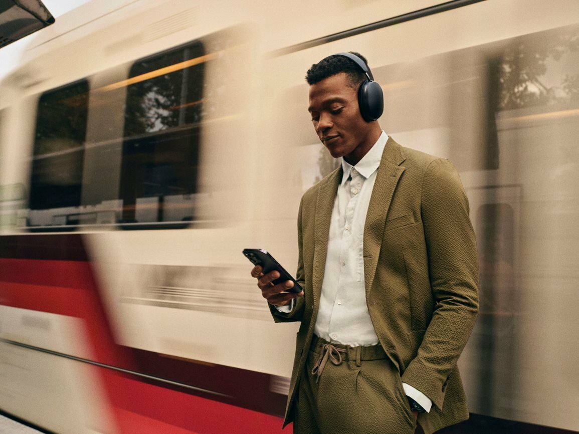 a male wearing the sonos ace stands on a train platform as the train whizzes by behind him