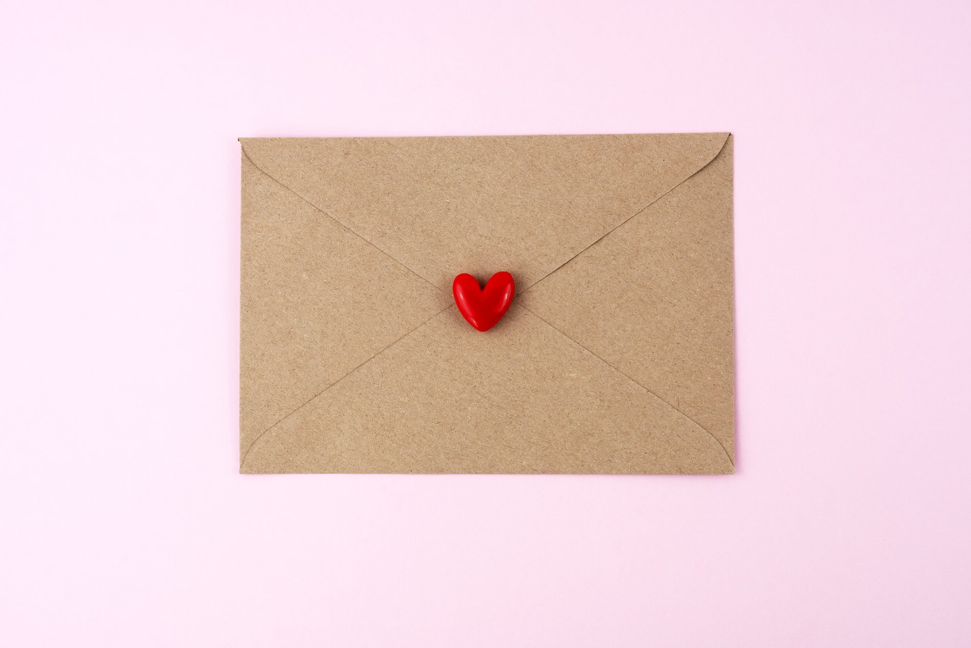 A brown envelope with a red heart on top of it, on a pale pink background. 