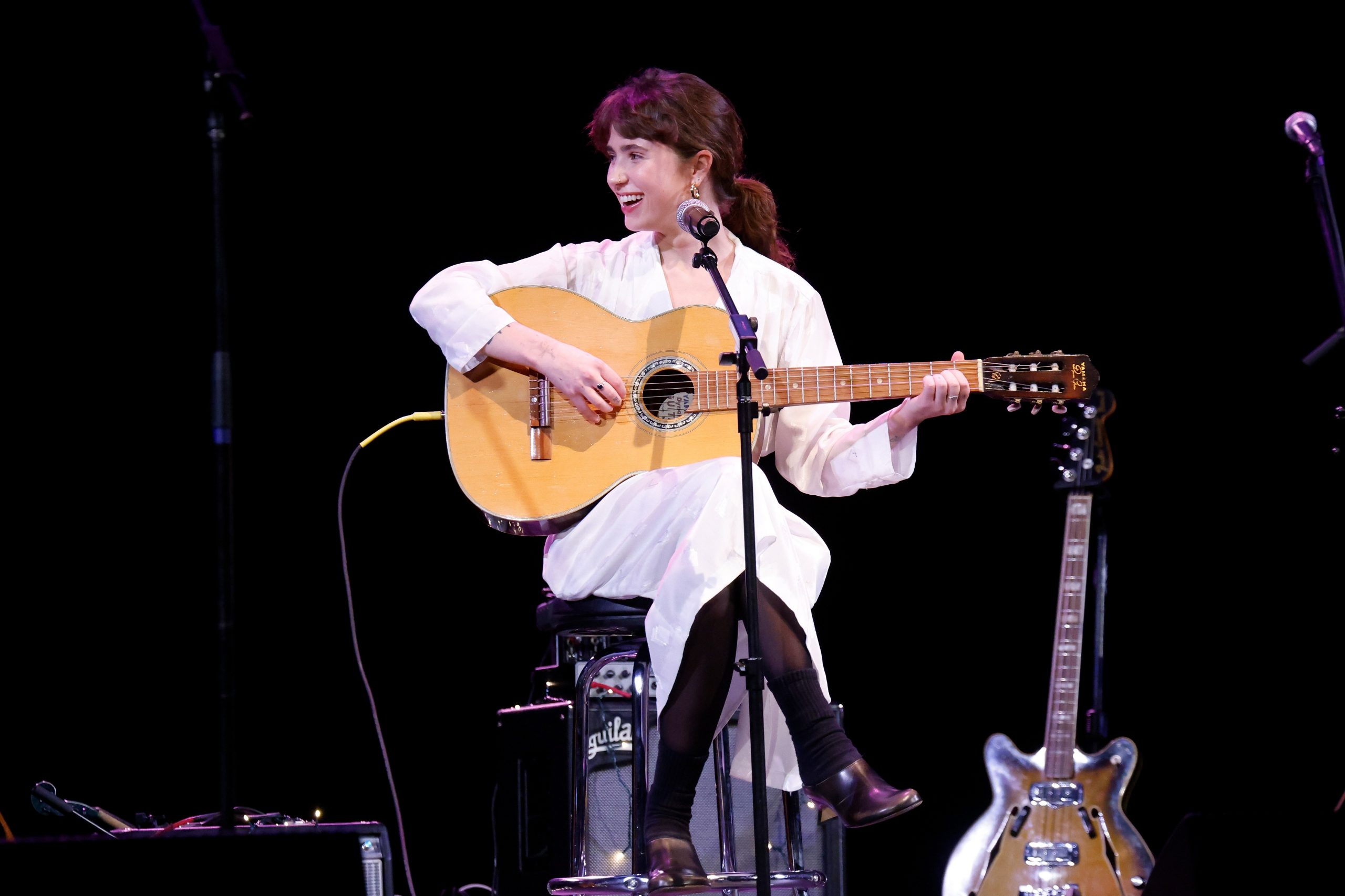 Clairo smiling as she plays the guitar on stage.