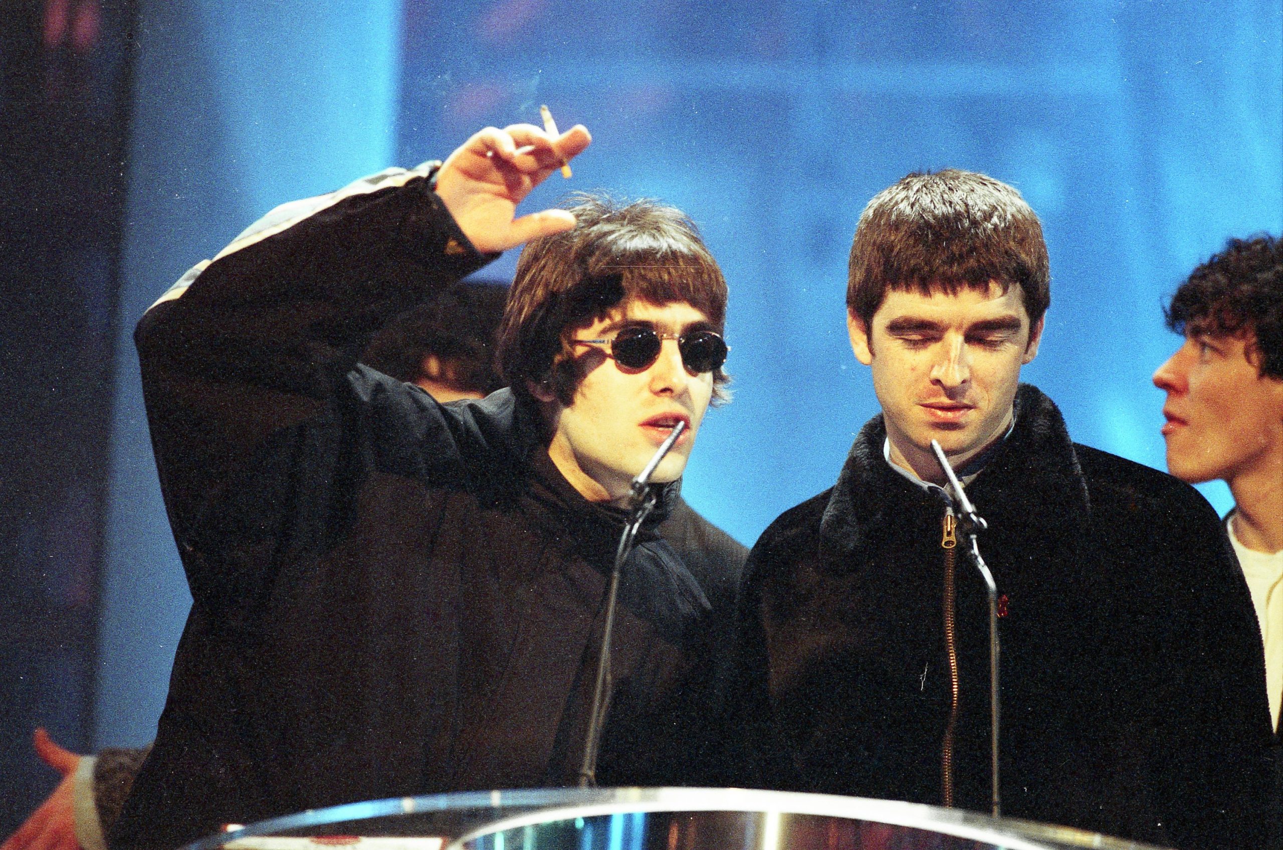 liam and noel gallagher on stage at an award ceremony. liam is gesturing with a cigarette in his hand, noel is looking downward.