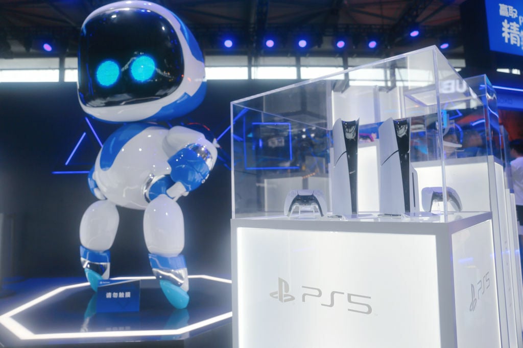 Two PS5 consoles in a glass display case next to a large Astro Bot statue