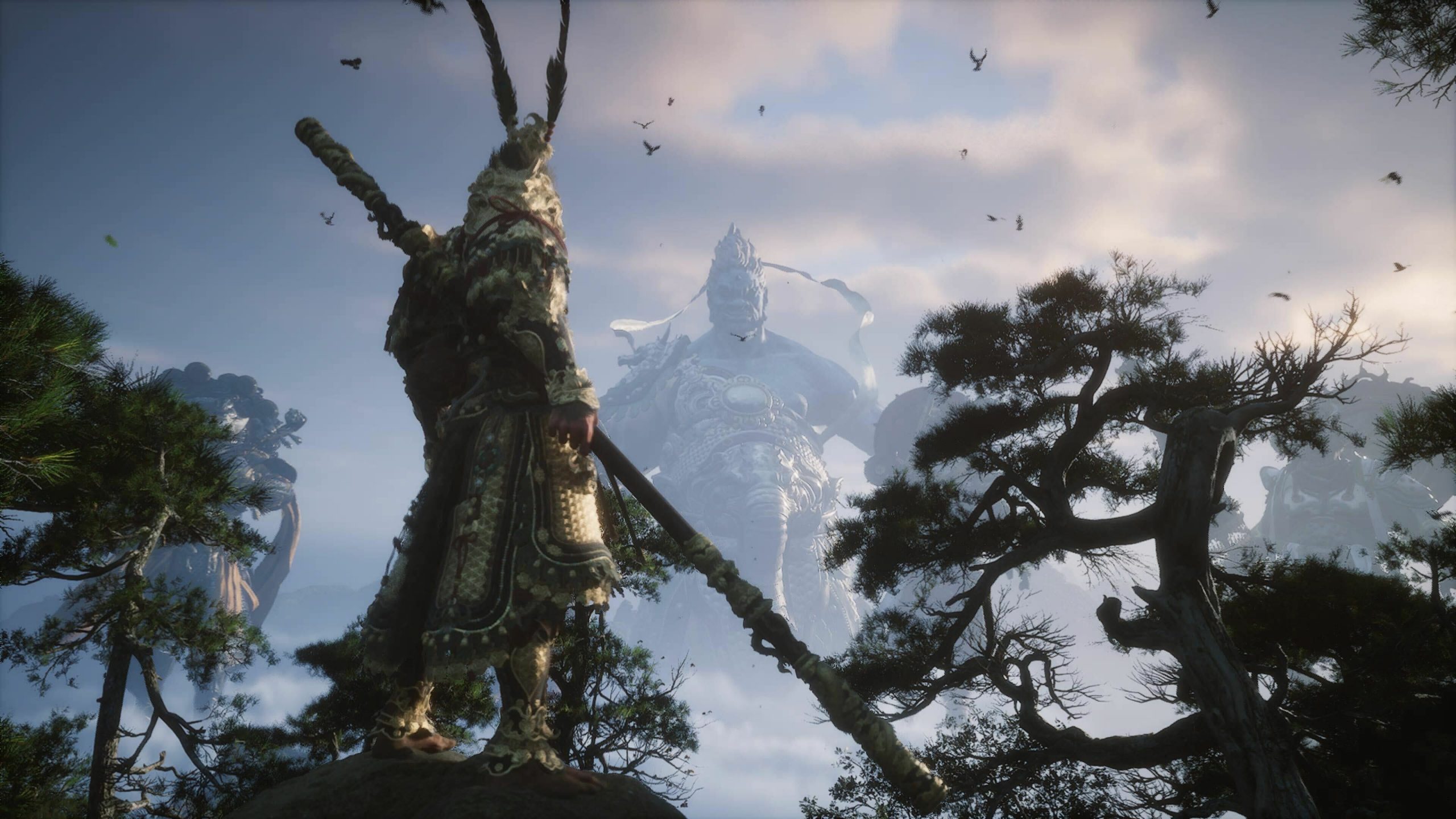 A warrior in ornate armor stands on a rocky cliff, holding a large staff, facing a colossal stone statue of a deity in the distance, surrounded by mist and trees. The scene is set against a cloudy sky with birds flying overhead.