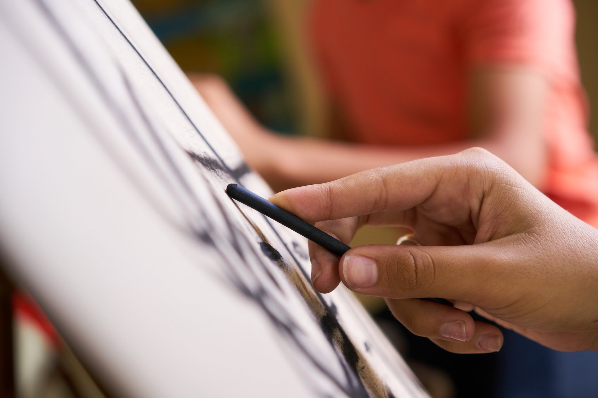 A close-up of a person's hand holding charcoal as they draw on an easel at an art class. 