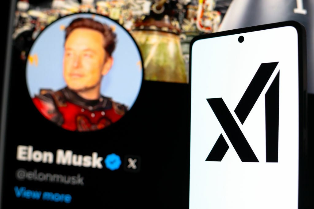 the xAI logo seen displayed on a smartphone with the official profile of Elon Musk, of the social network X in the background