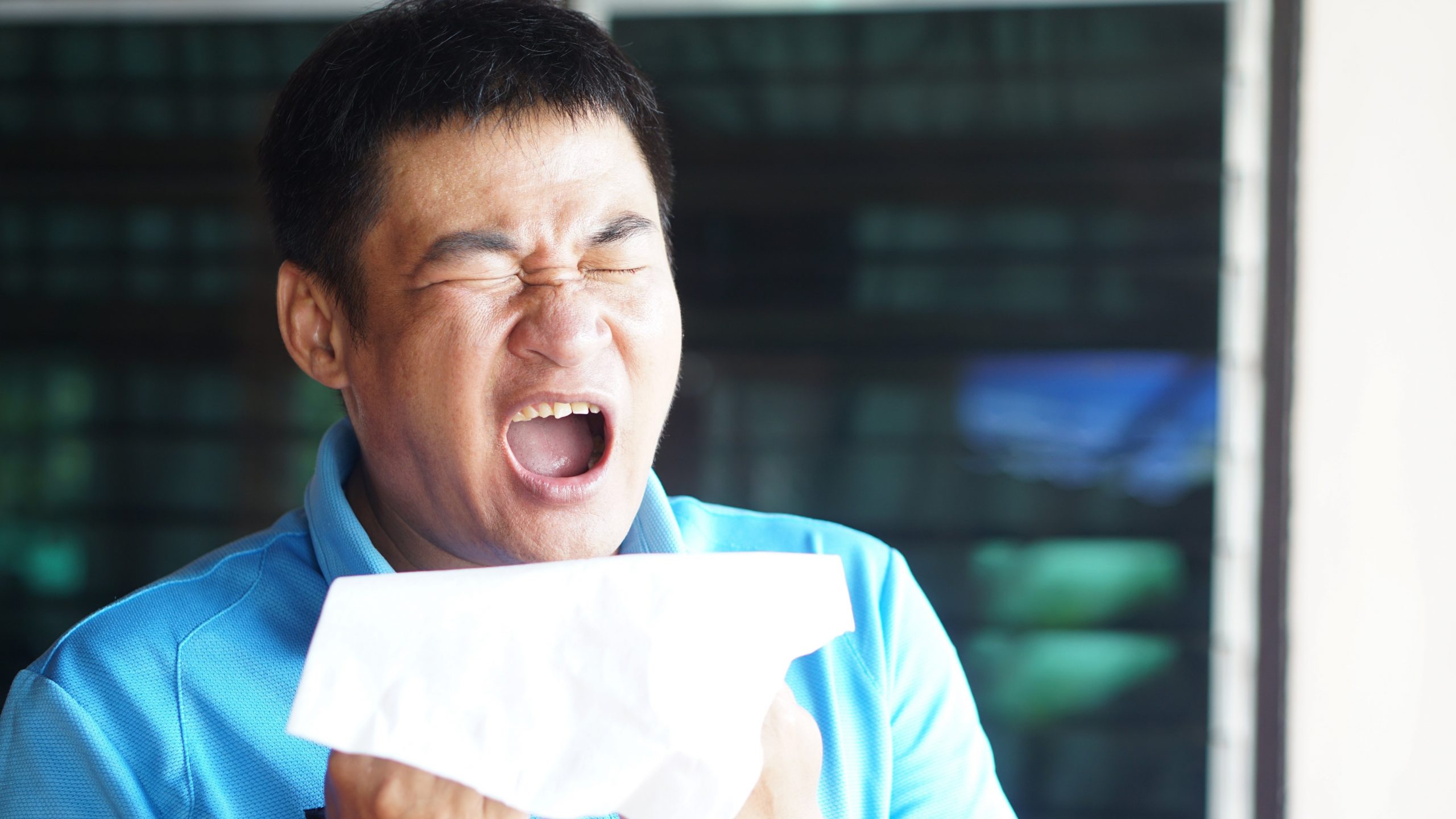 Man in a blue shirt sneezing while holding a tissue