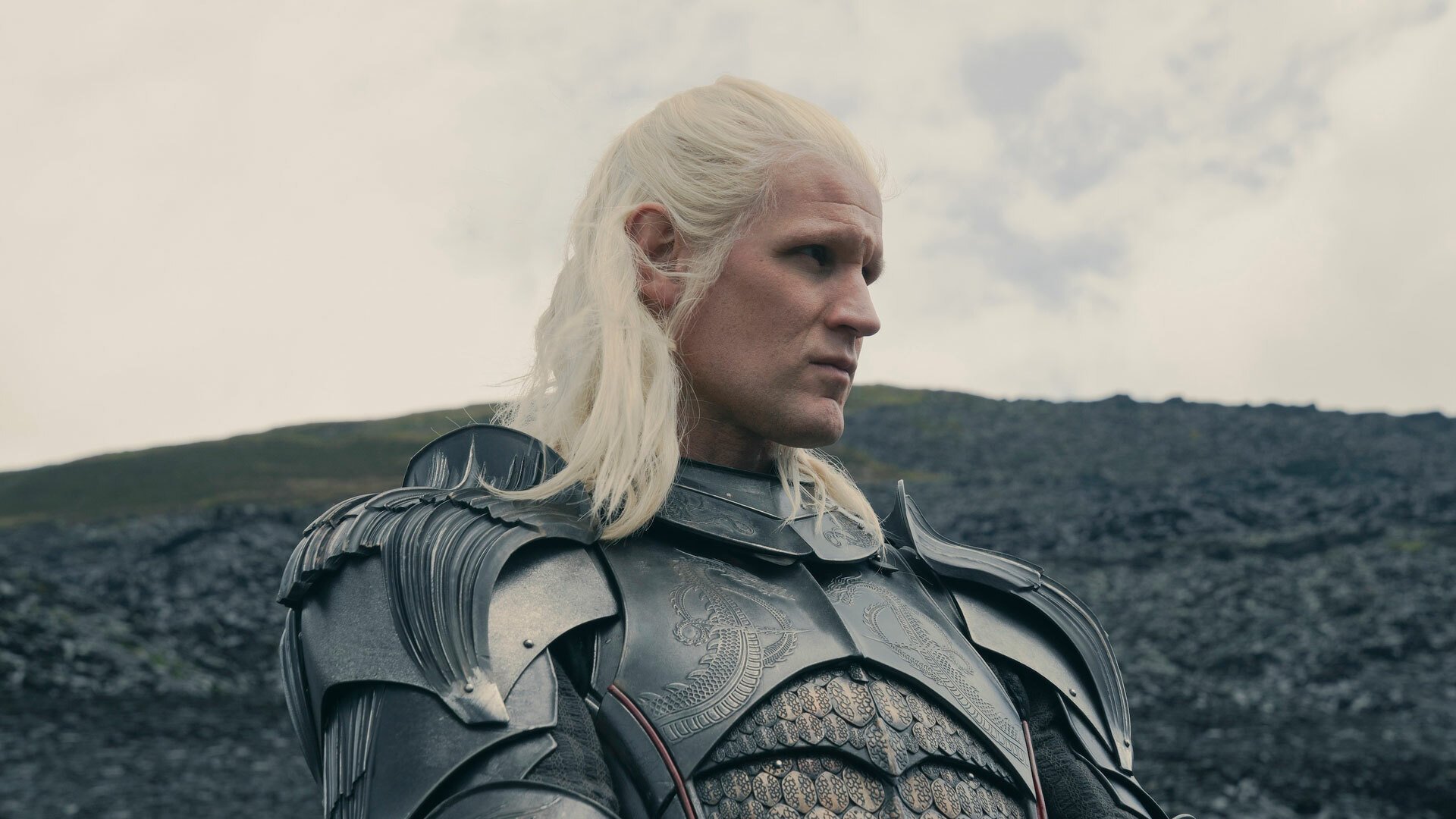 A man with long blonde hair in armour stands on a hillside.