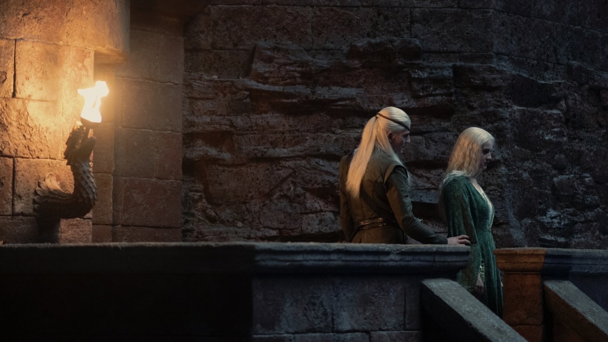 Aemond Targaryen grabs Helaena Targaryen's arm; the two stand on a torchlit parapet.