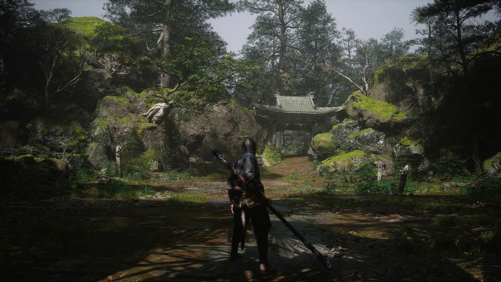 A warrior holding a staff stands in a lush forest clearing, facing a moss-covered rock formation with a traditional, weathered gate nestled among the trees. The scene is tranquil, with sunlight filtering through the dense foliage and reflecting off the shallow water on the ground.