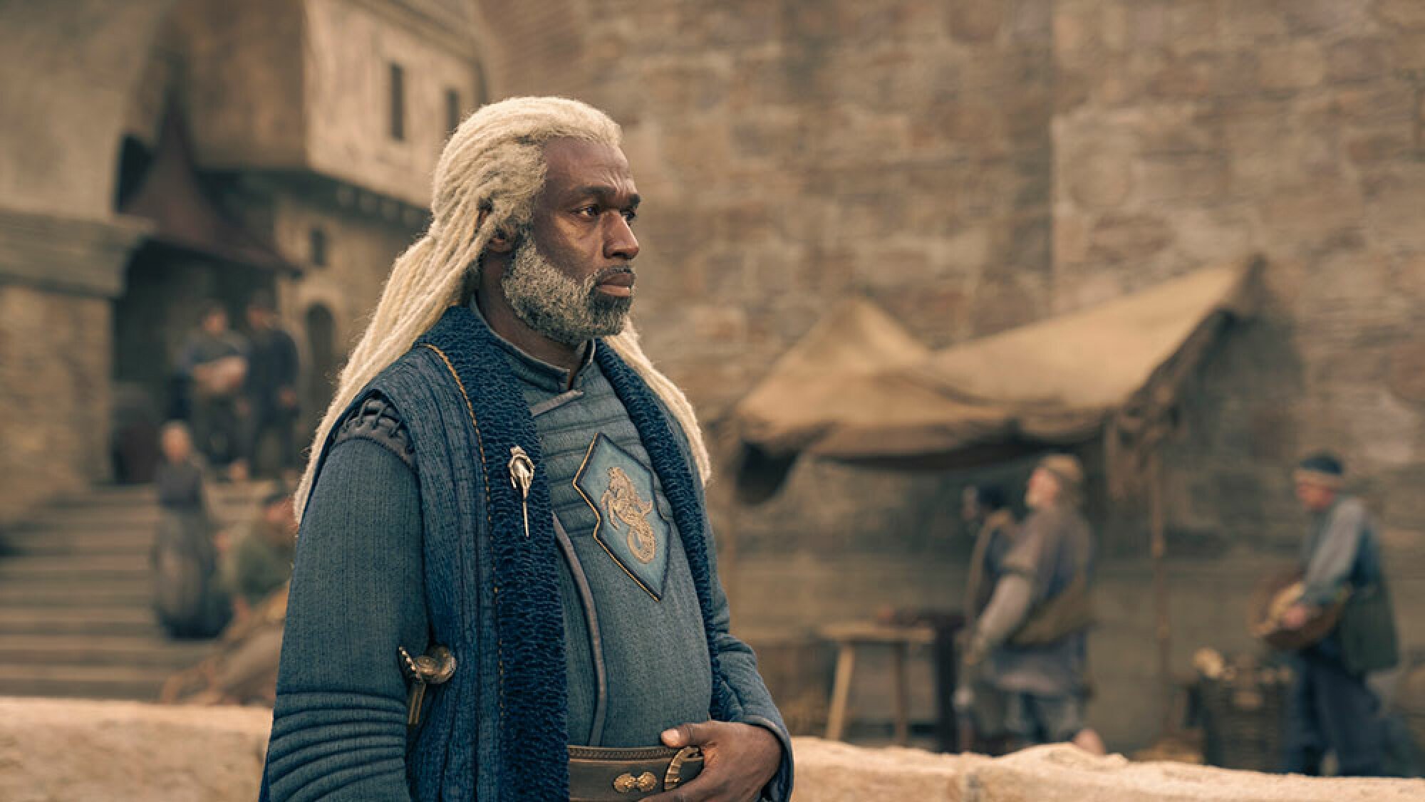 A man in medieval clothing with long white hair stands outside in a harbour.