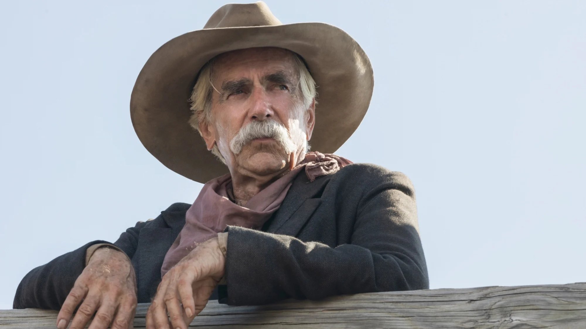 An old man with a mustache and cowboy hat leans on a wooden fence. 