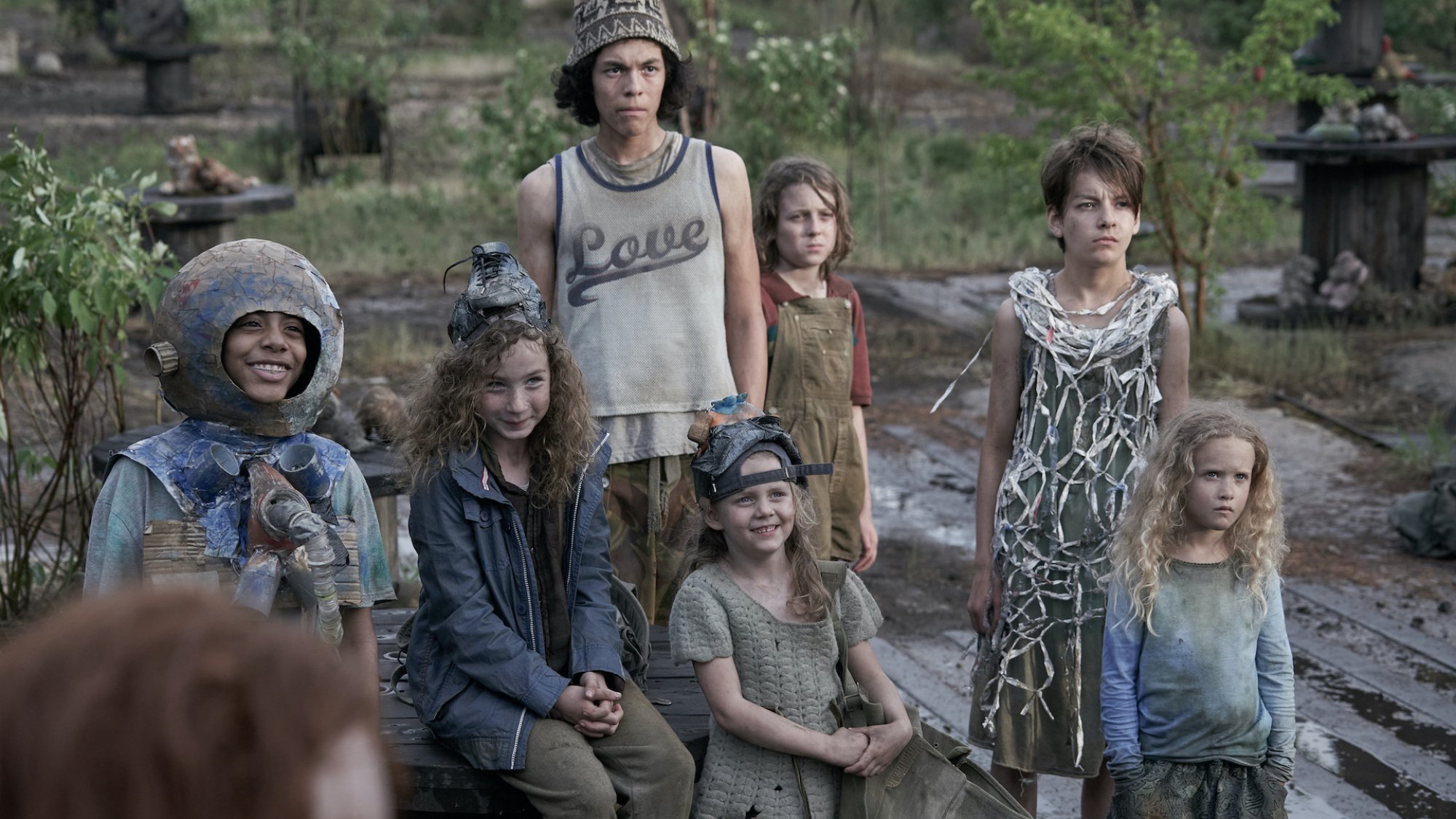 A group of kids wearing tattered clothing sit around in a desolate space. 