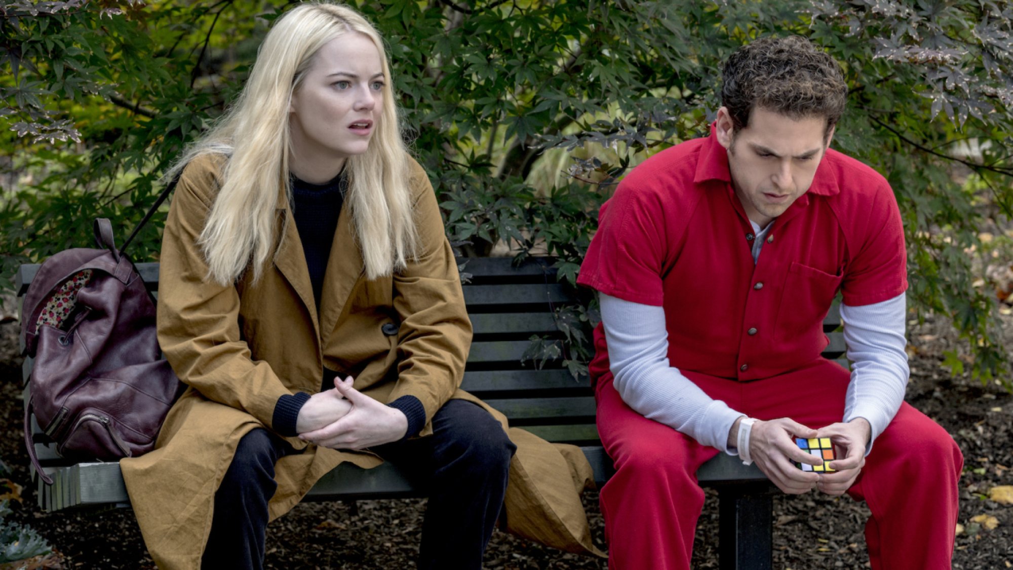 A woman wearing a yellow coat sits on a bench with a man wearing a red jumpsuit. 