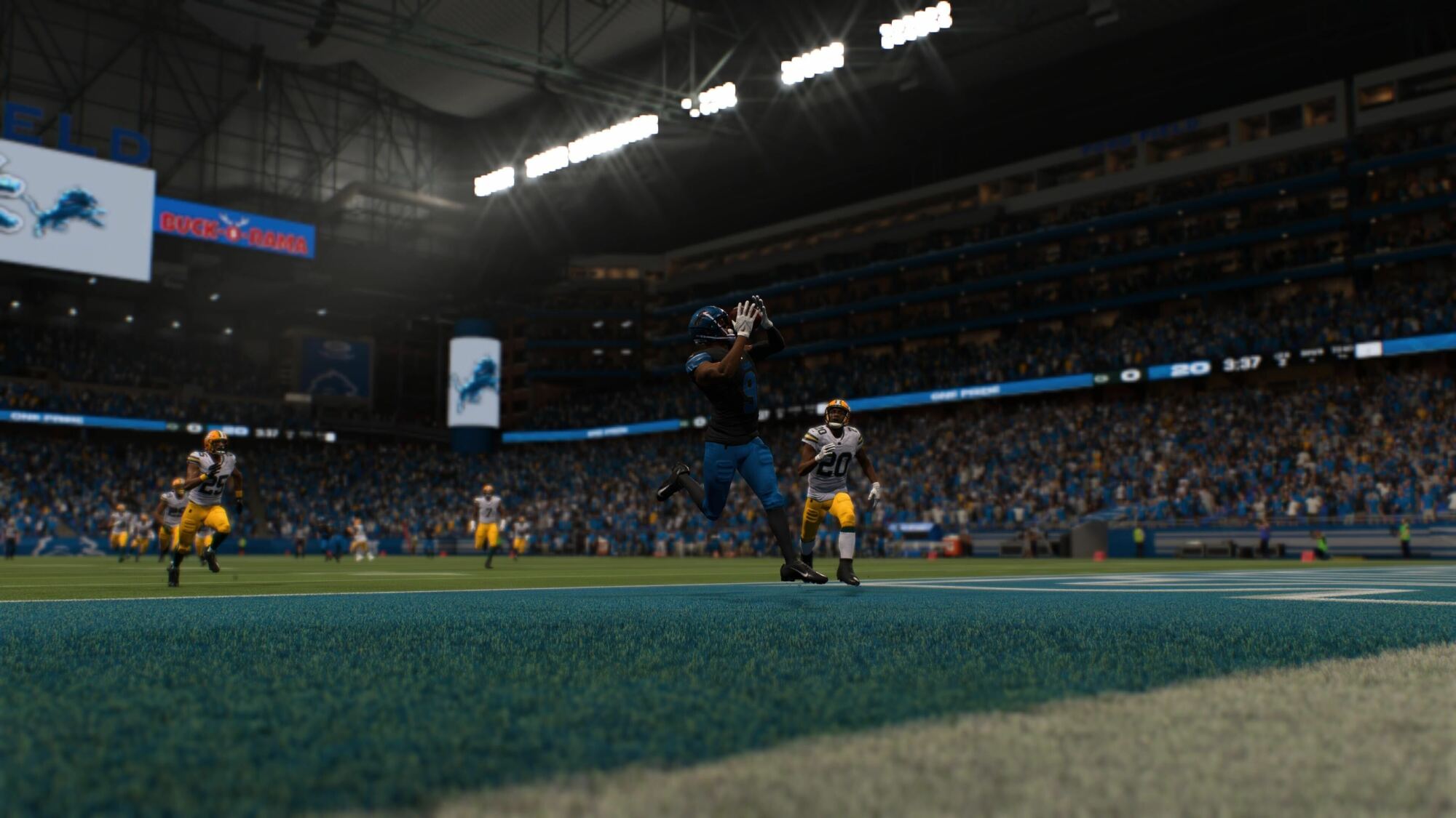 A player wearing a blue and black uniform is catching a football in the end zone, scoring a touchdown. He is closely followed by a defender in a yellow and green uniform. The stadium is filled with spectators, and the goalpost is visible in the background, along with the word "LIONS" written on the sideline, indicating that the game is being played at the Detroit Lions' home stadium. The crowd and environment suggest an intense moment during the game.