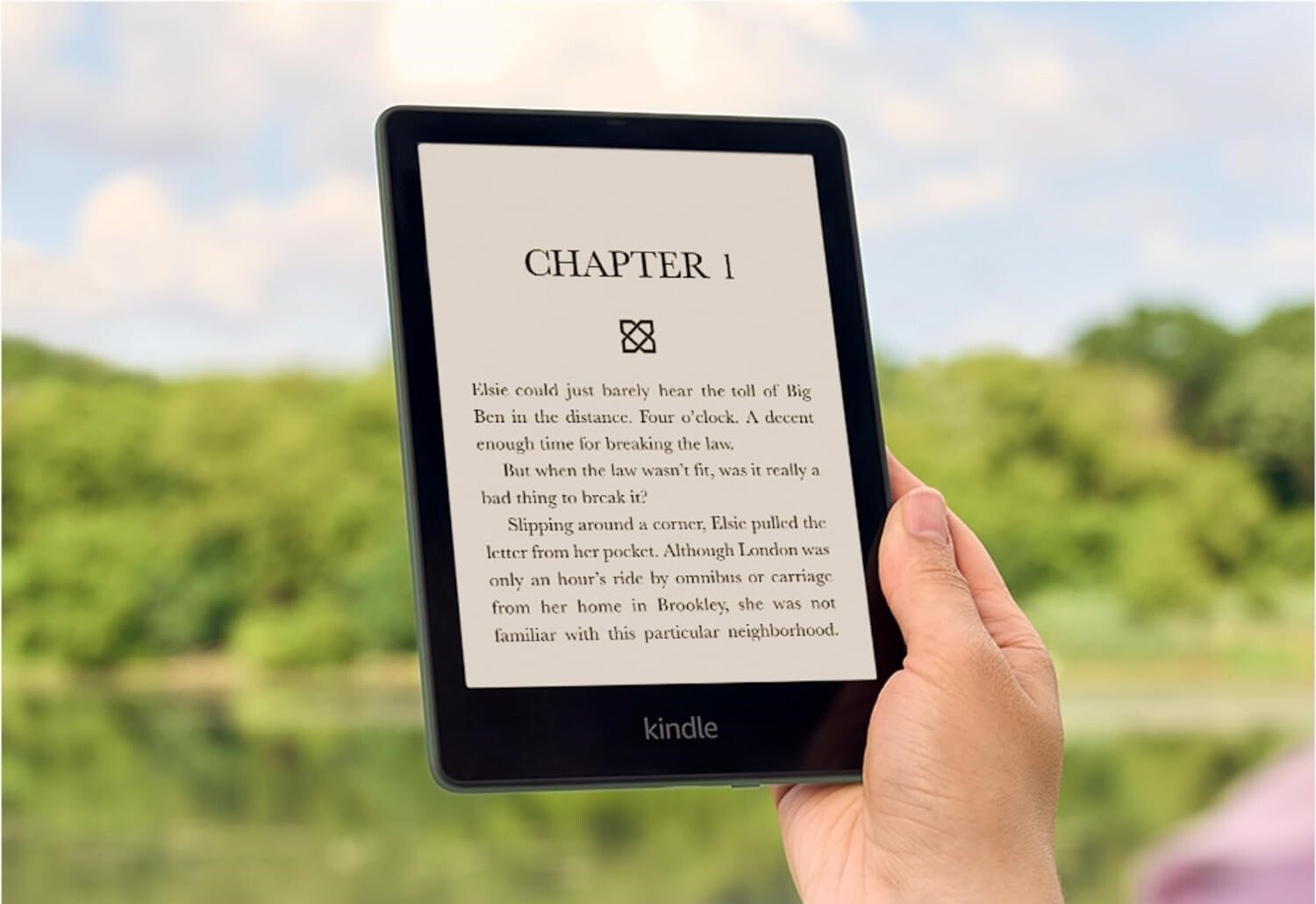 a person holds the amazon kindle in their right hand. The background has a forest scene with trees.