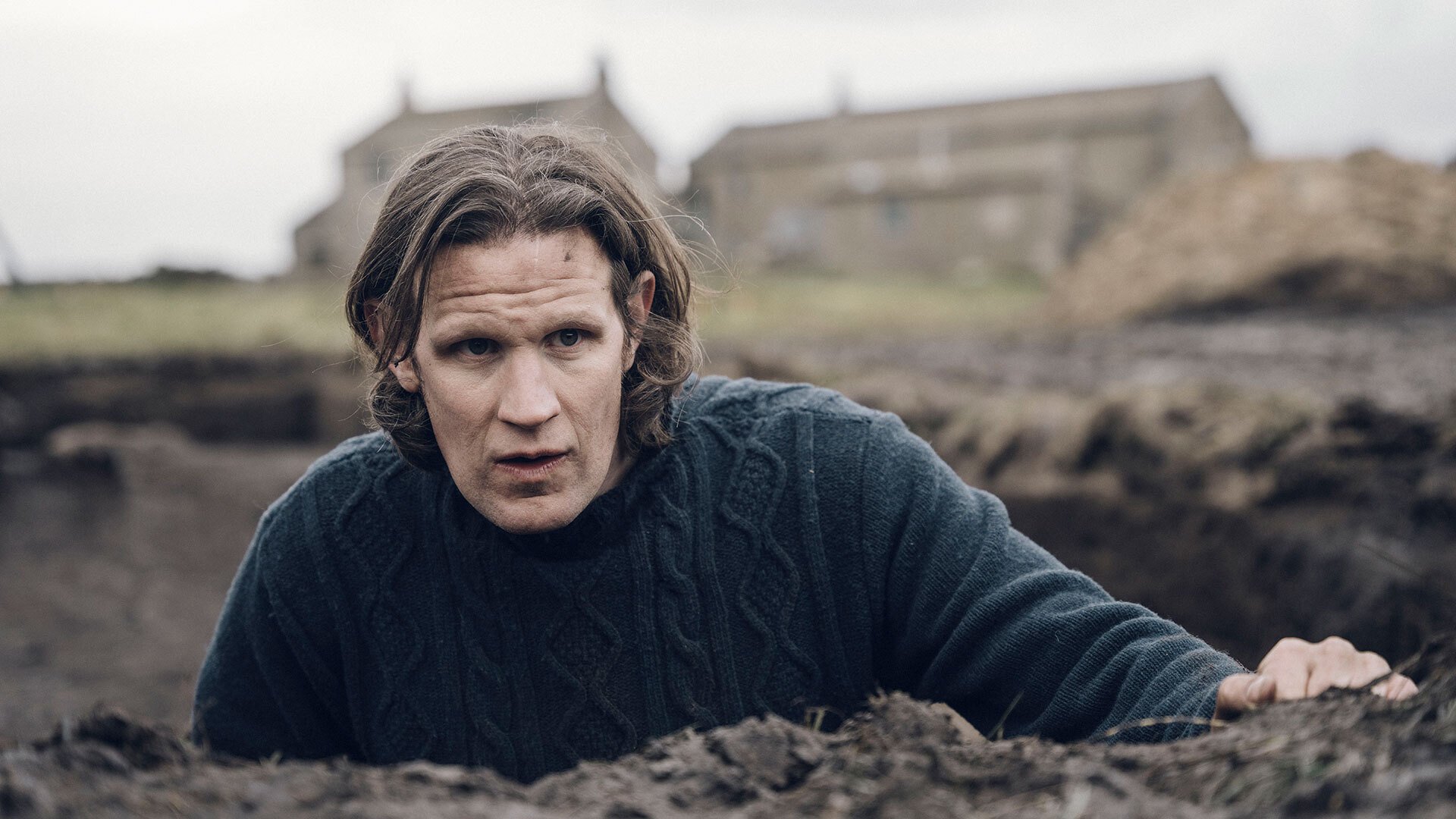 A man standing in a trench in the ground in the countryside looks worried. A farmhouse is visible in the background.