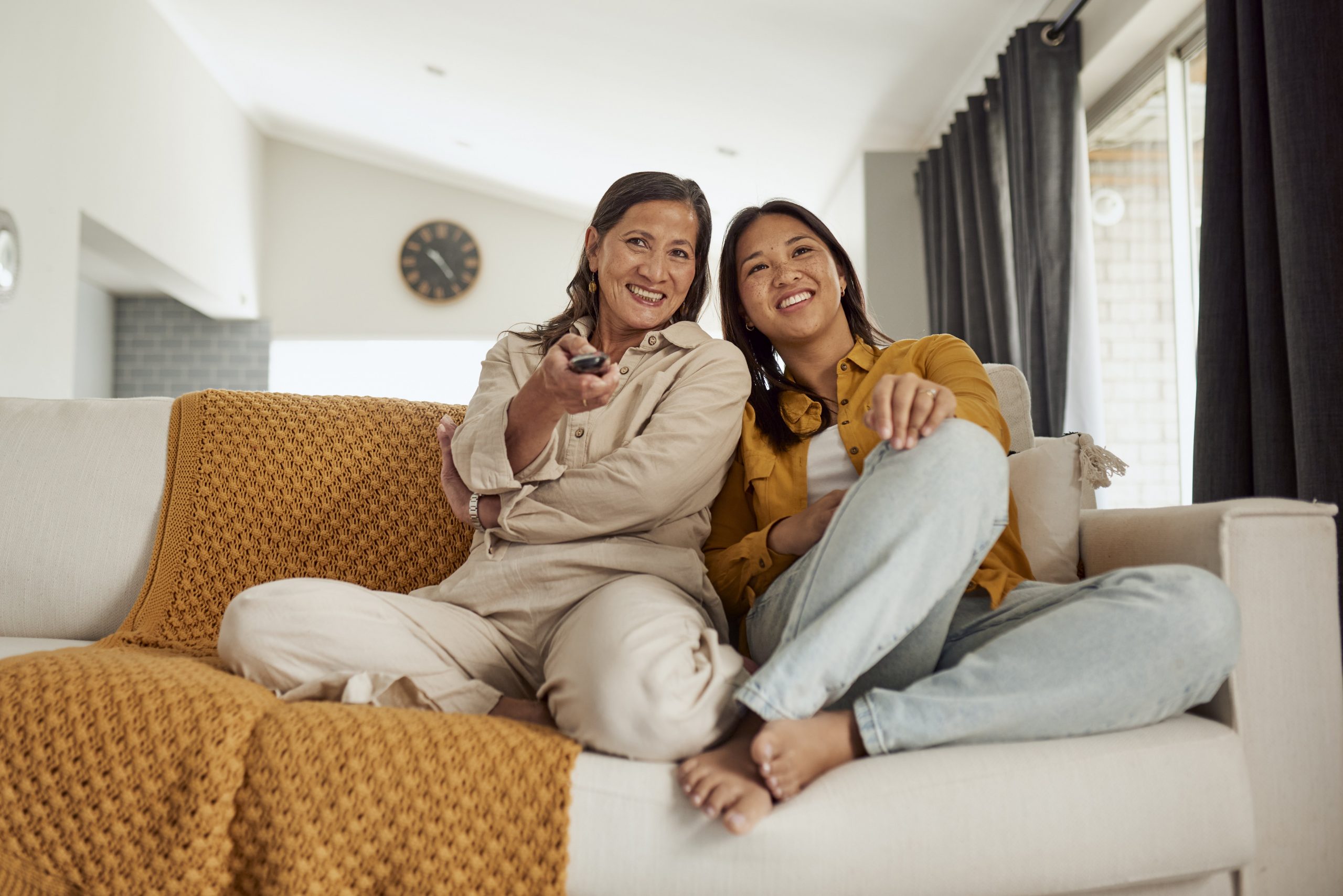 two people sit on a cream-colored couch while one of them hold a TV remote and both smile