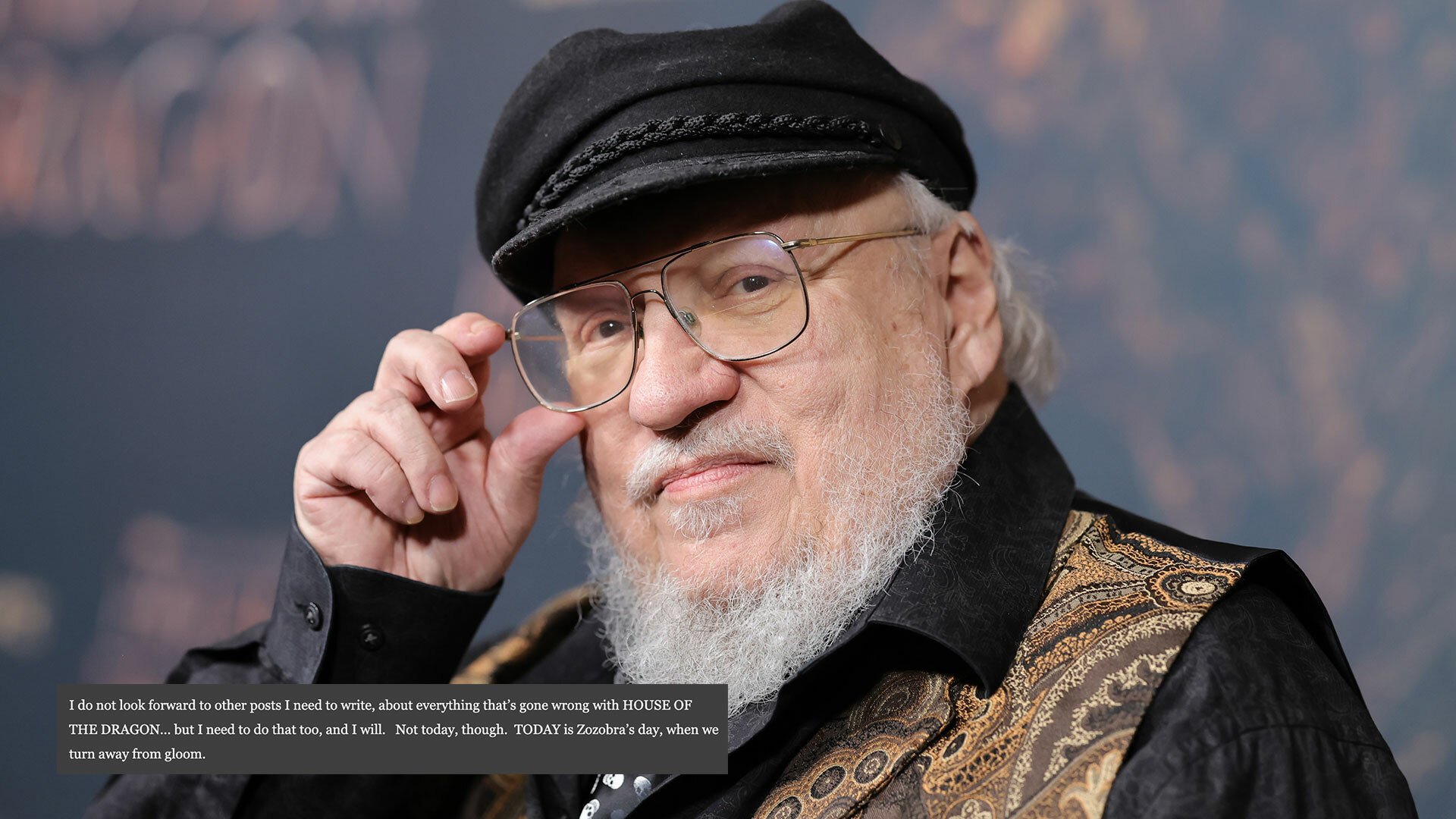 A close-up of a man with glasses, a white beard and a hat standing on a red carpet.