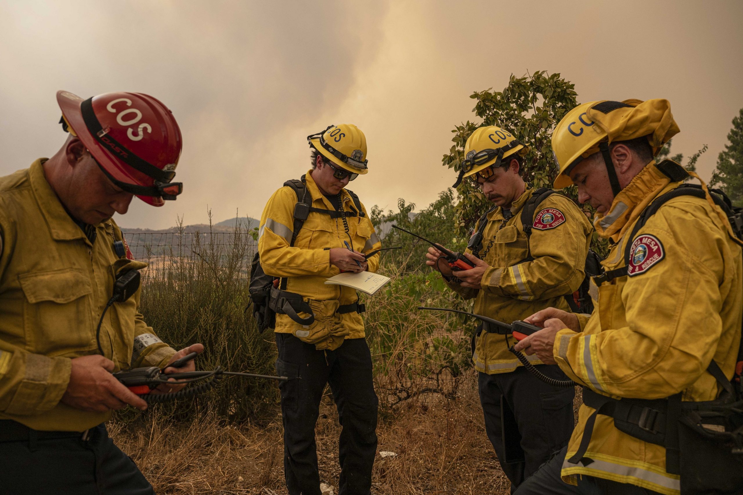 Four firefighters stand in a circle looking down at radios and notepads. 