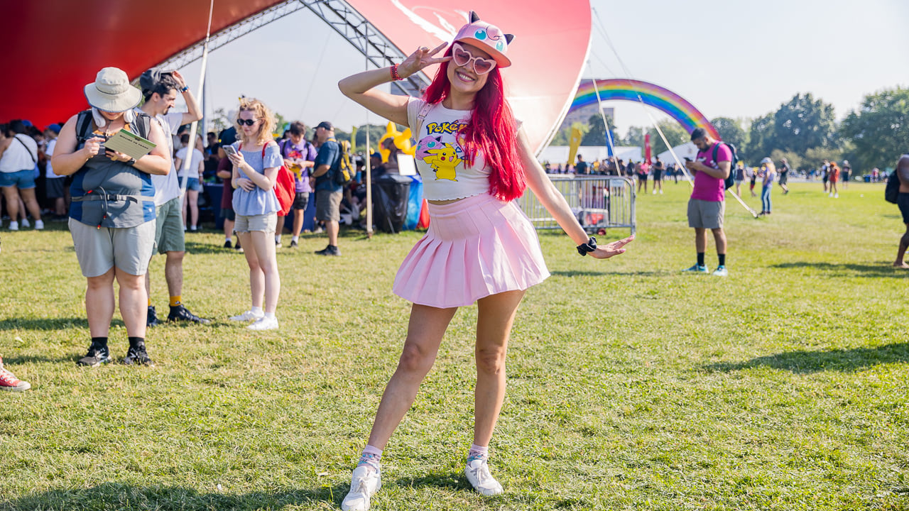 A women in a light pink pleated skirt, white sneakers, and cap. She has red hair and is making a peace sign with her left 