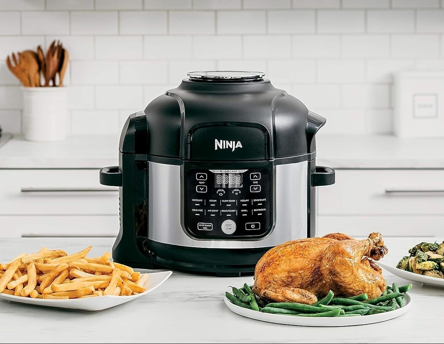 a ninja foodi air fryer and pressure cooker combo sits on a kitchen counter with several dishes of food in front of it including a roast chicken, french fries, and brussel sprouts